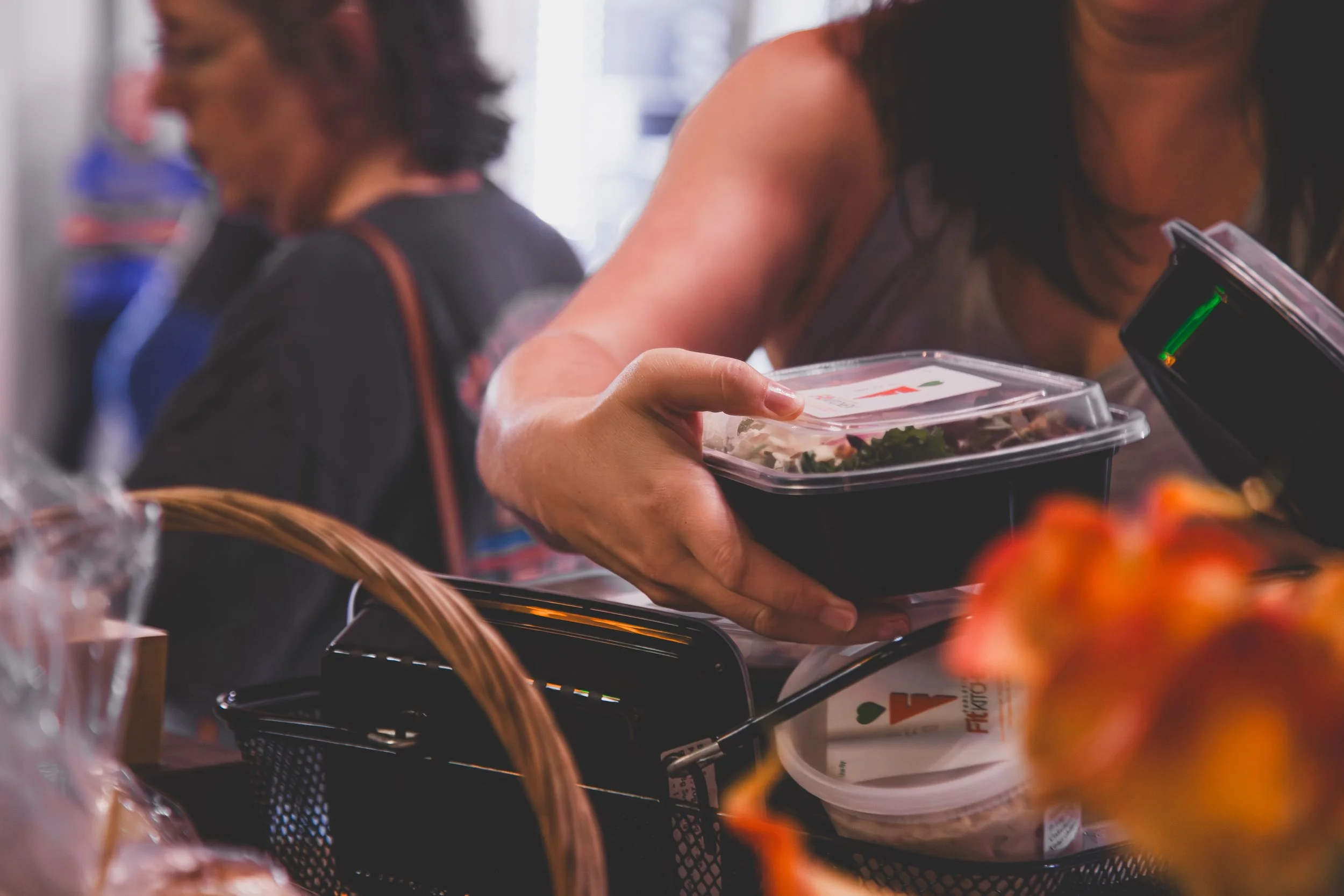 A woman reaching for a takeout salad container in a grocery store or market, with a basket and other food items visible.