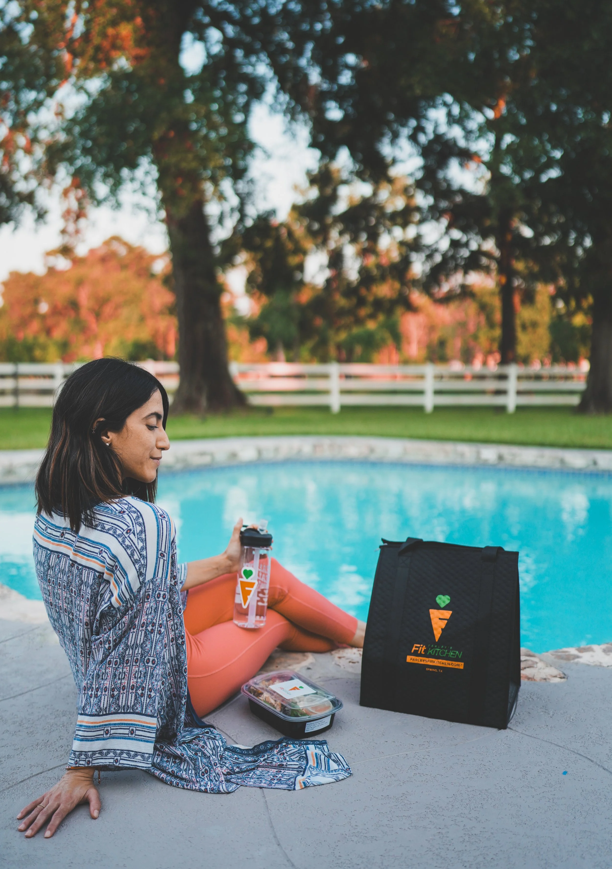 A woman sitting by a swimming pool with a black grocery bag and a water bottle, about to eat a lunch container. The scene is outdoors with trees and a white fence in the background during sunset.
