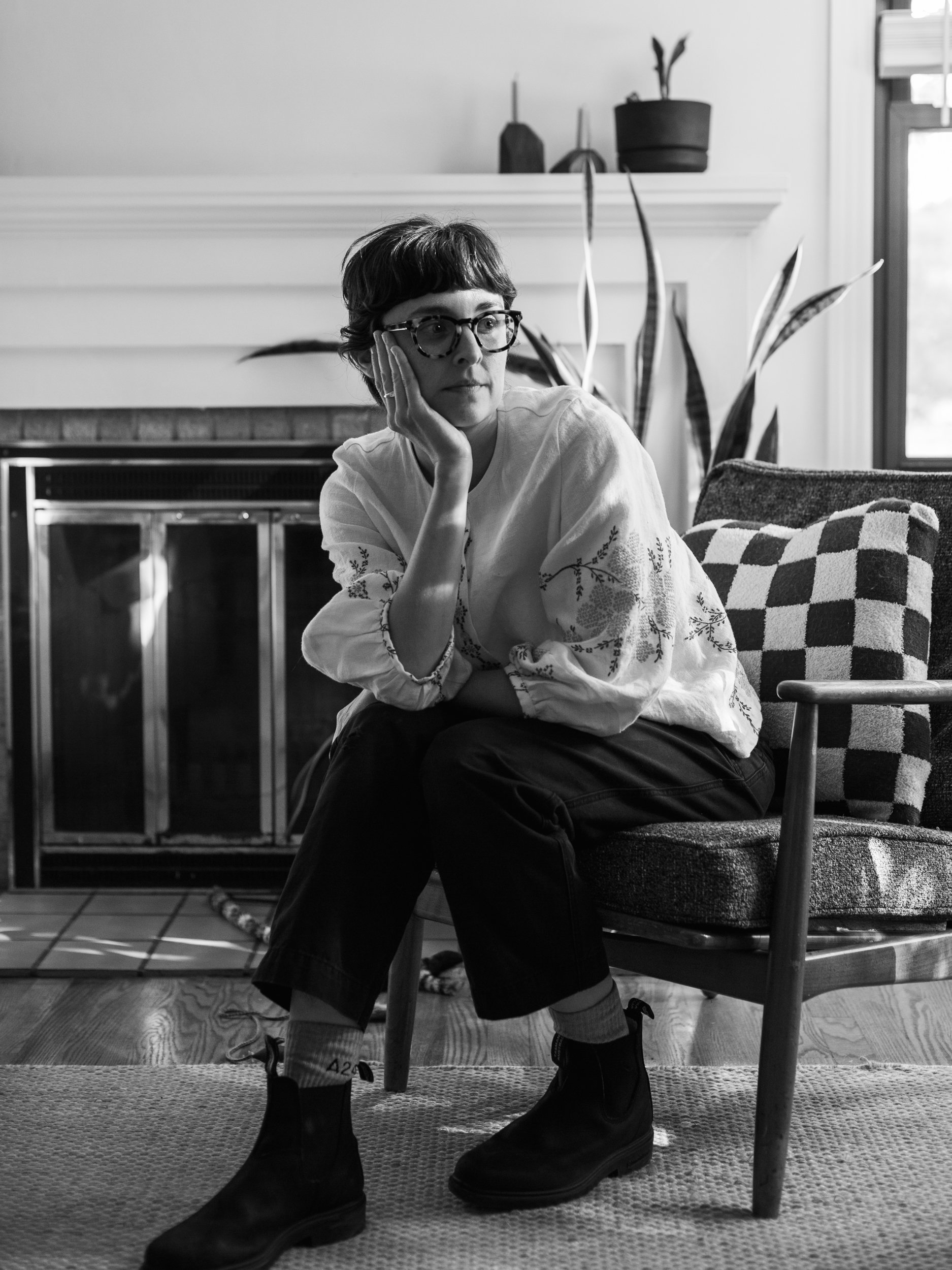 A woman sitting on a chair with her chin resting on her hand, wearing glasses and a white embroidered blouse, in a living room with a fireplace, plants, and a checkered pillow.