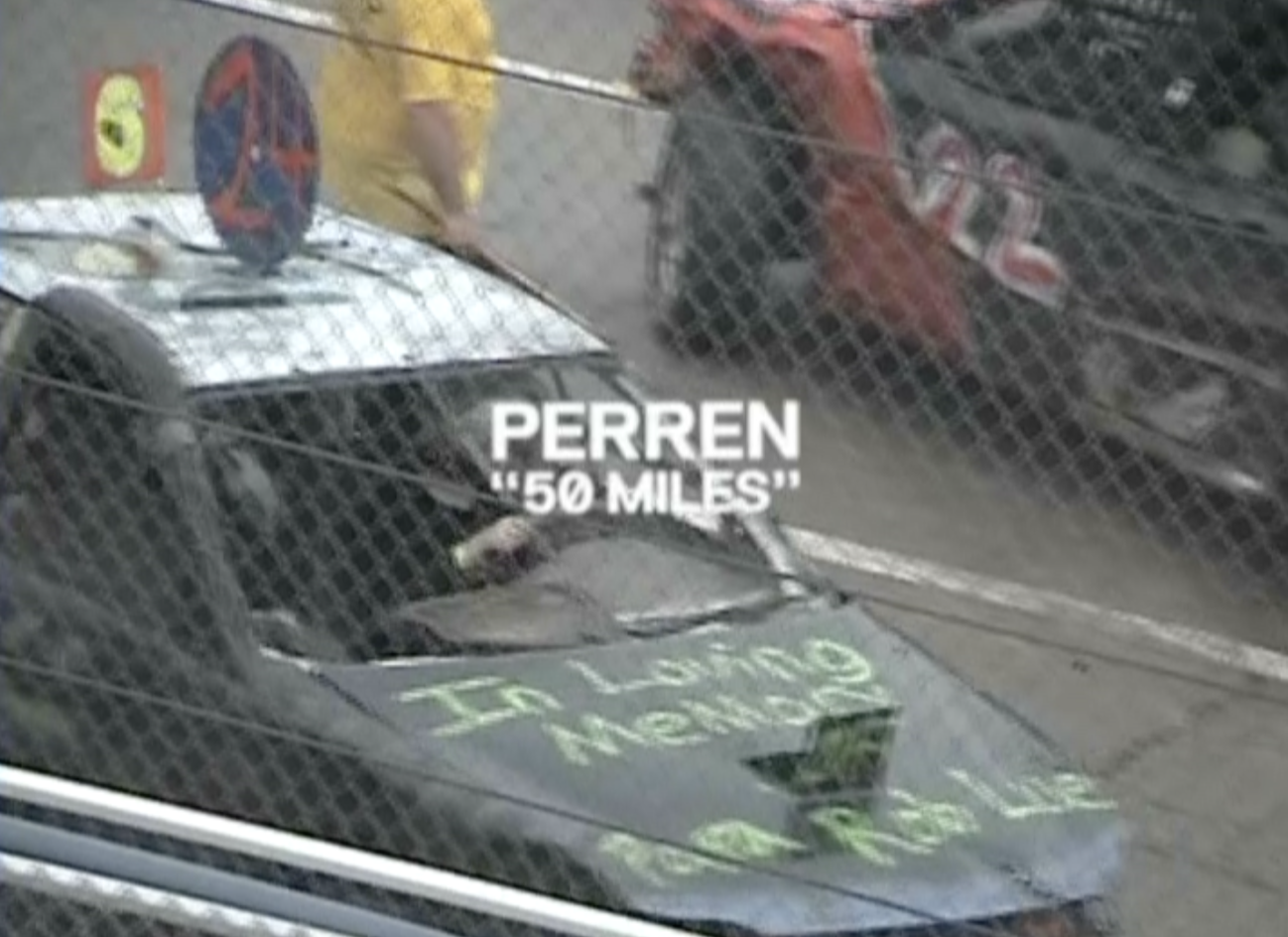A car with a sign on the hood reading "In the Name of Nature" and a message in the windshield protesting Perren, who is 50 miles away, as seen through a chain-link fence.