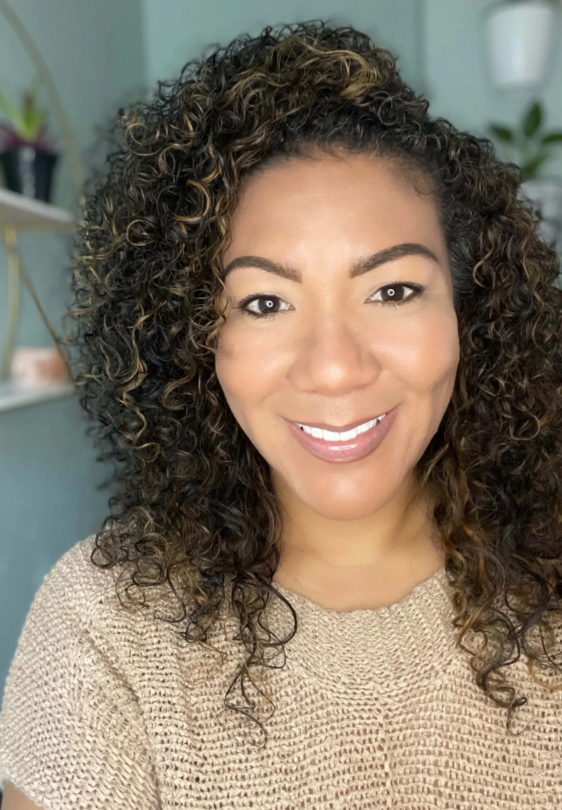 A woman with curly dark hair and light brown highlights, smiling indoors.
