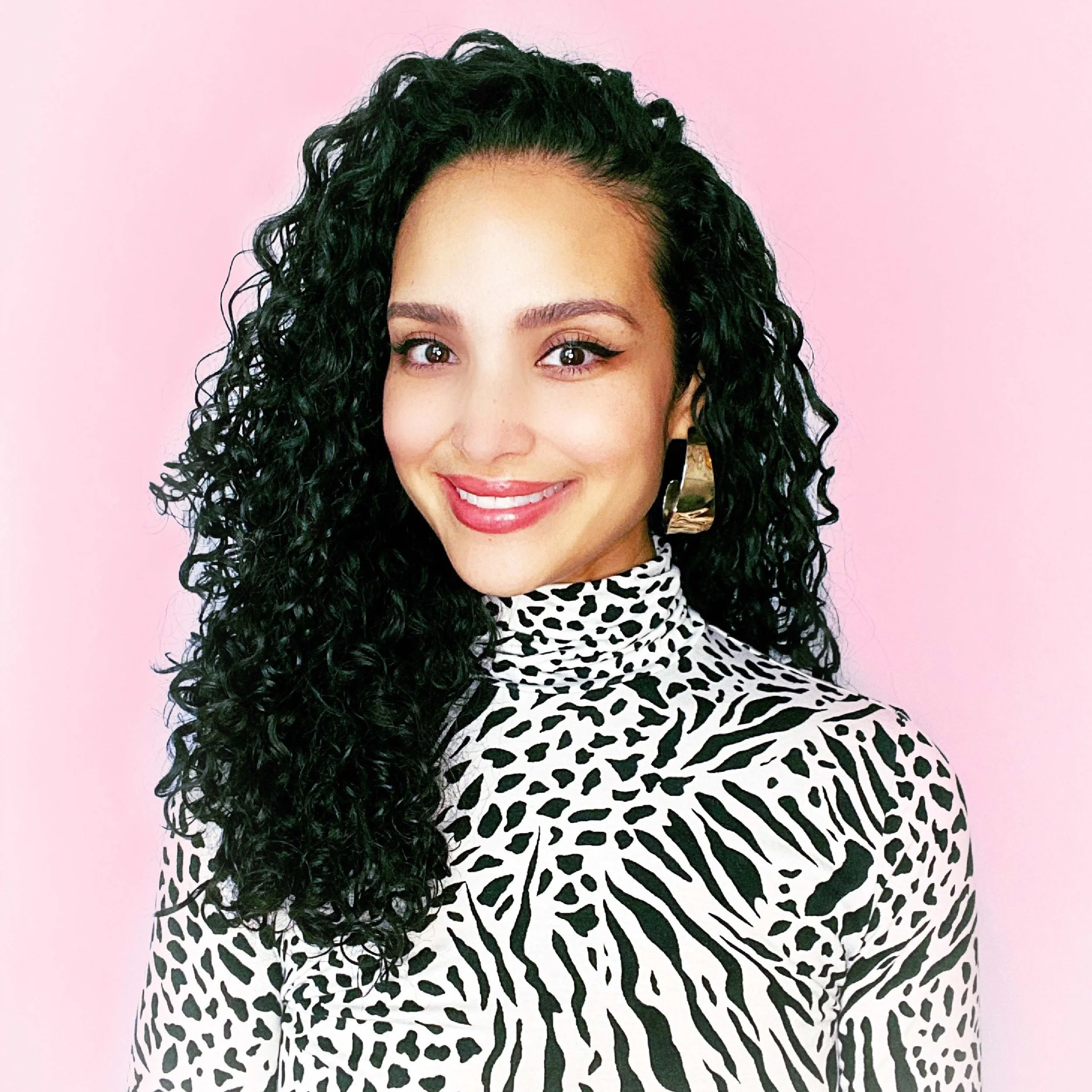 Portrait of a woman with long dark curly hair, wearing a black and white animal print turtleneck top and gold earrings, smiling against a pink background.