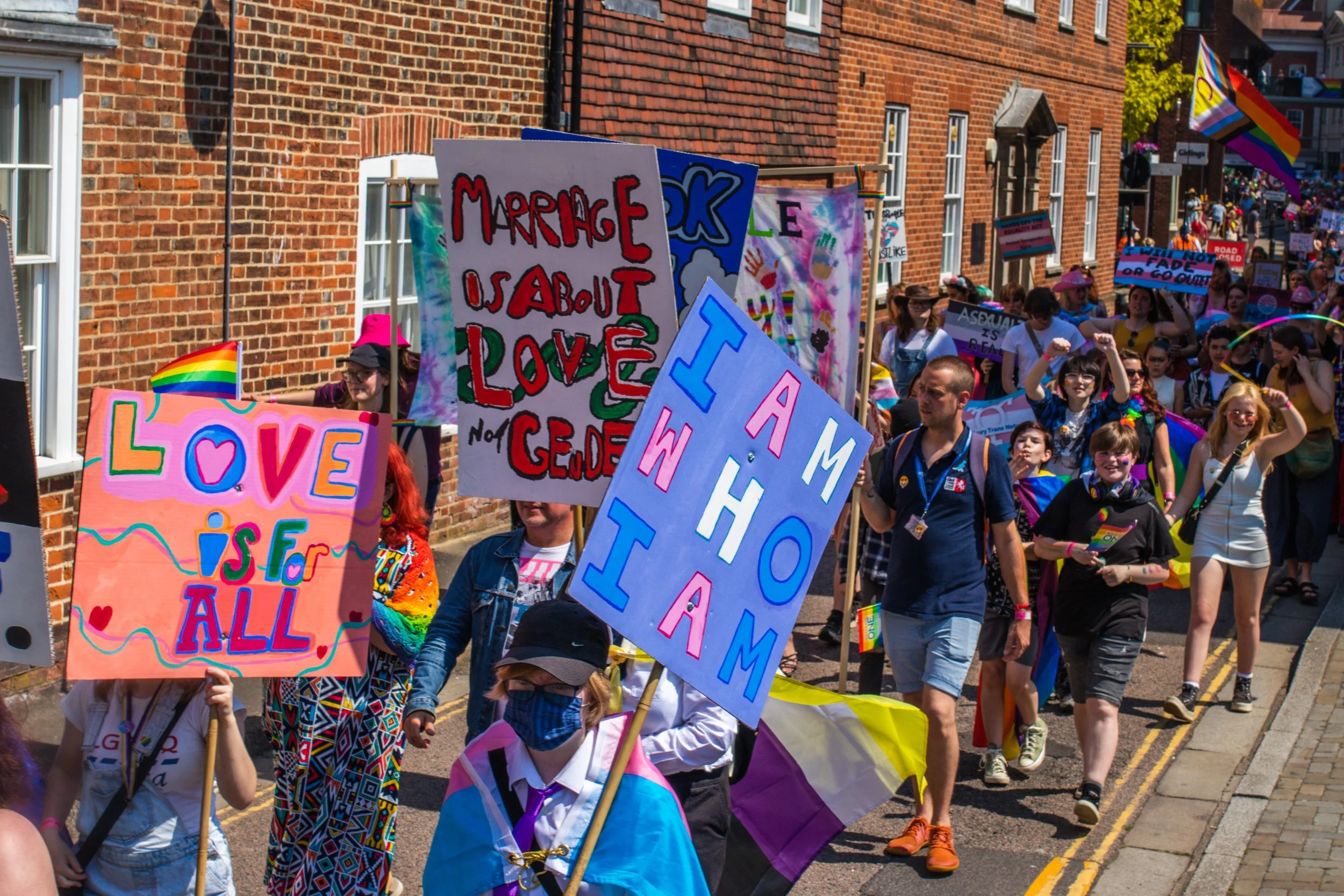 Photo showing groups in the Pride Canterbury Parade, with placards visible saying phrases like I Am Who I Am, and Love Is For All.