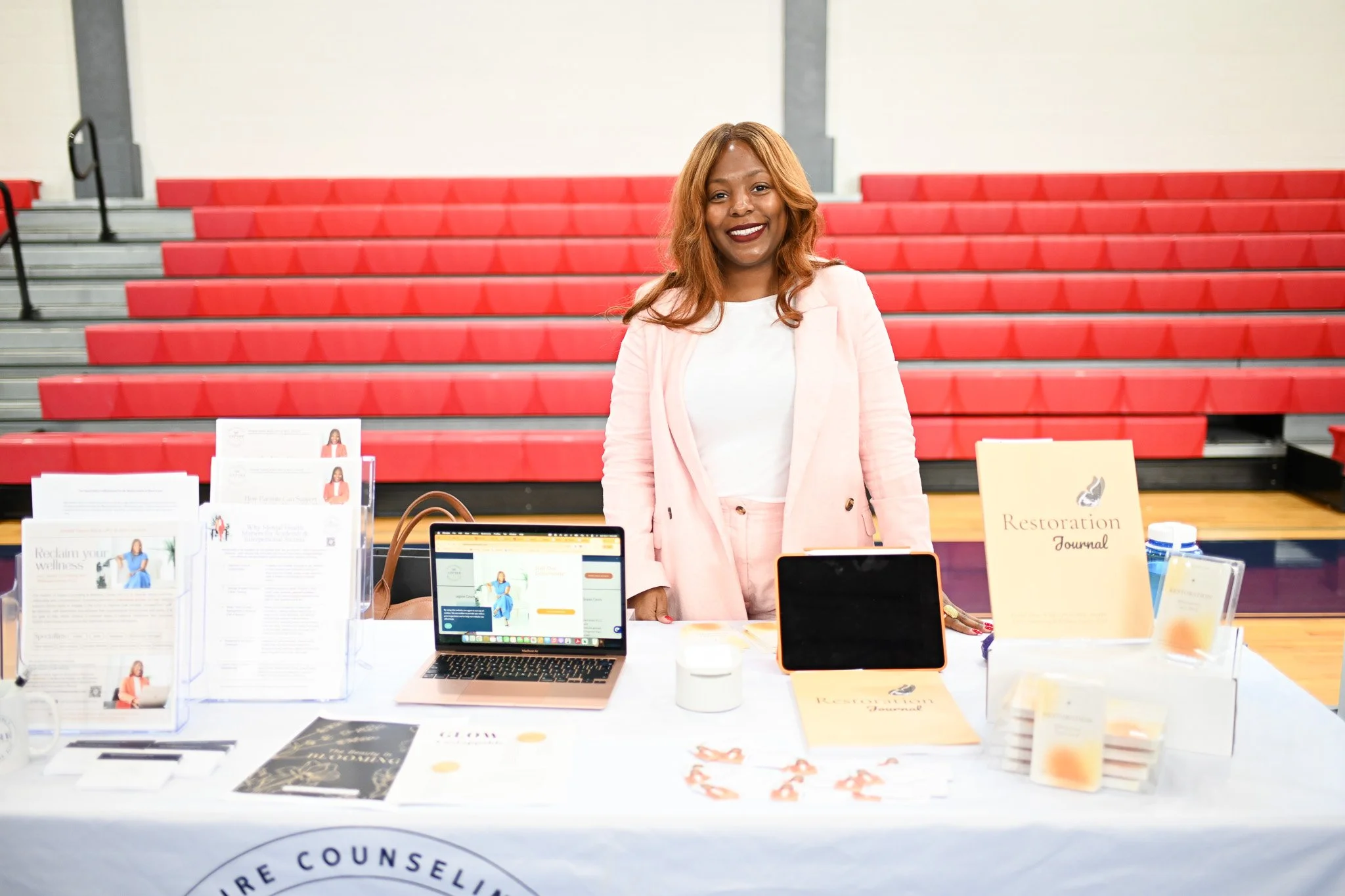 A woman with brown hair and a white shirt standing behind a table at an event, smiling, with informational brochures, a laptop, and a sign that reads 'Restoration Journal' on the table. A red bleacher background is visible.