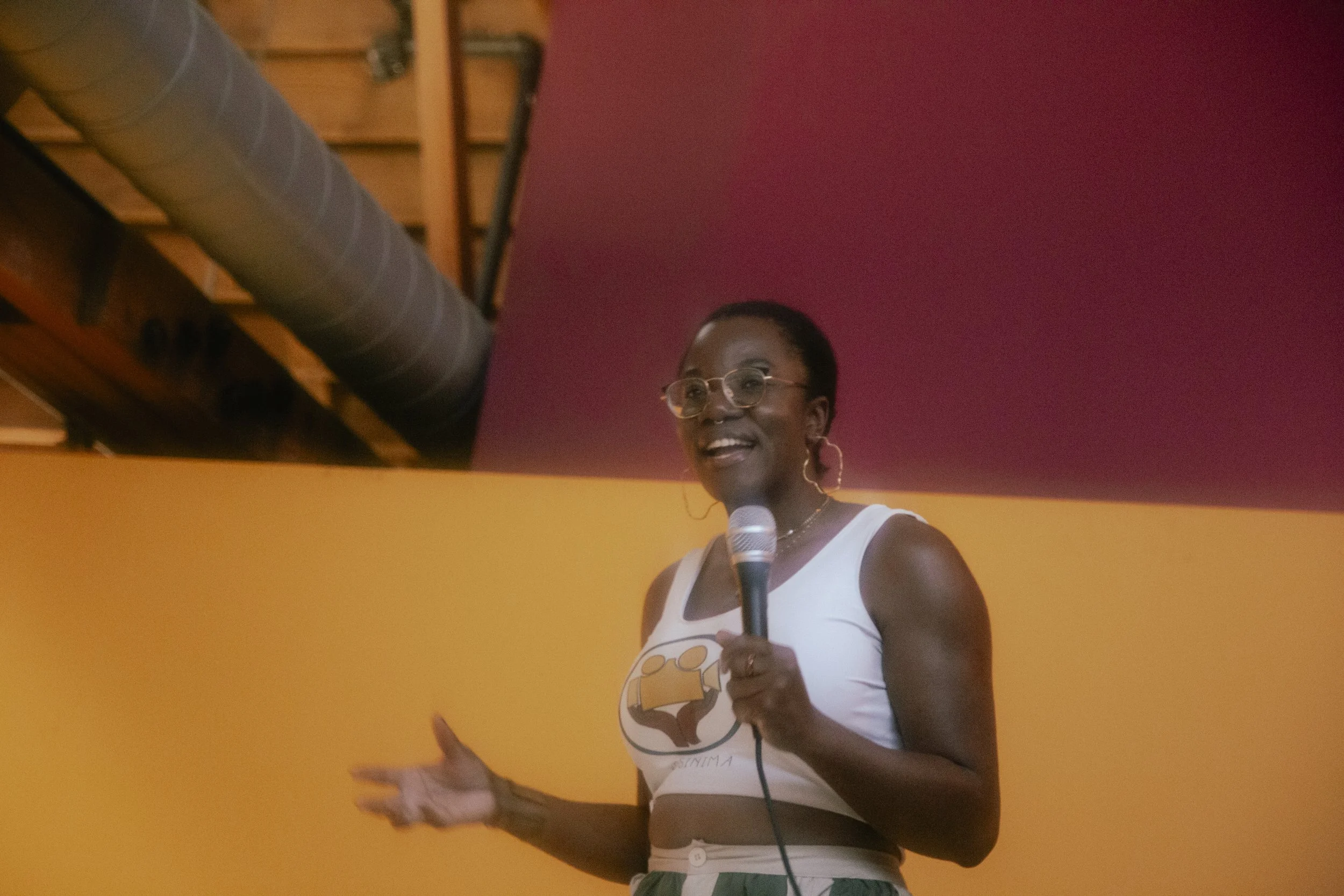 Oluwaseun Babalola, a young dark skinned Black woman wearling glasses and a white tank top with the kosinima inc logo, holding a microphone in front of a red and oranhe background.