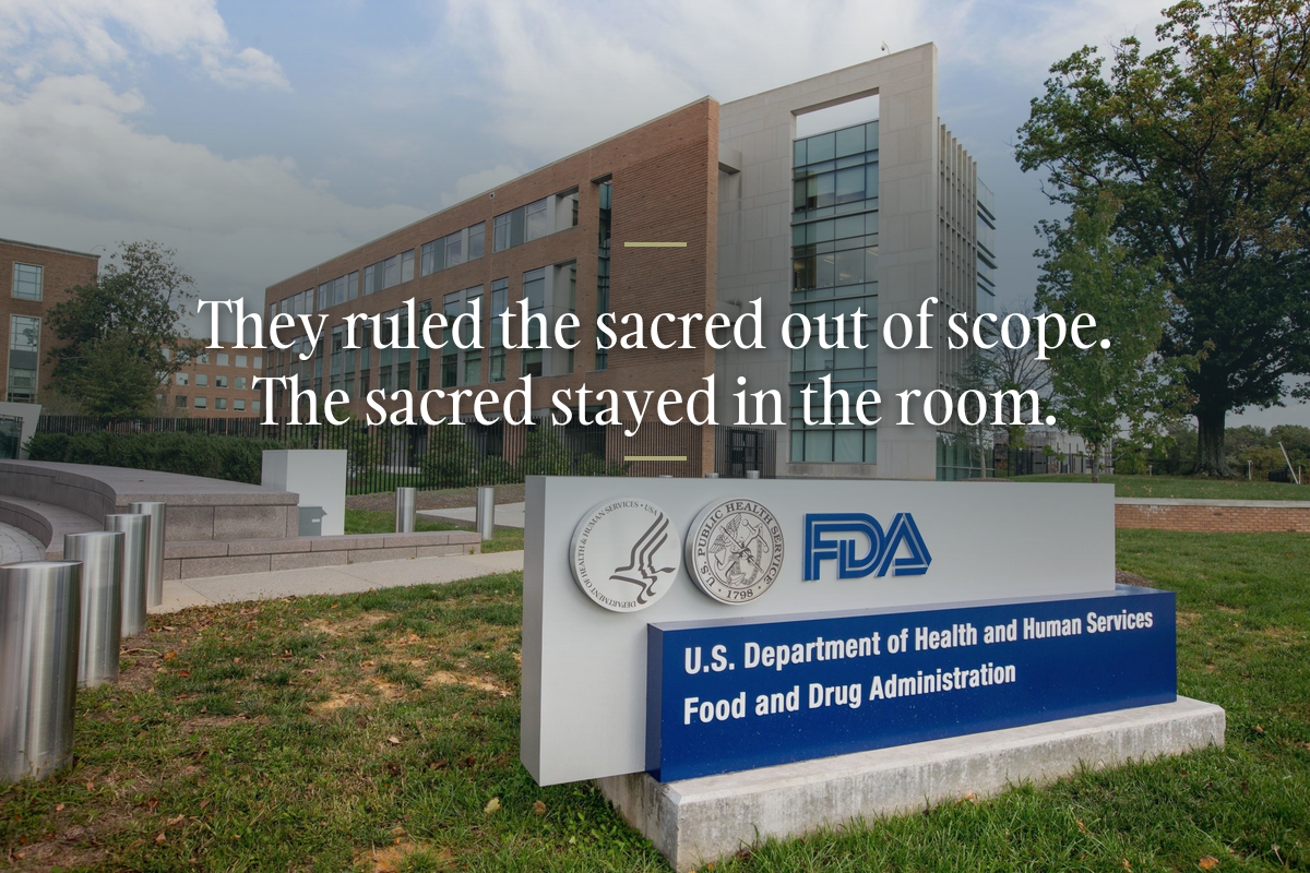 The FDA sign at the White Oak campus in Silver Spring, Maryland, brick building and oaks behind it, with the line in white serif: They ruled the sacred out of scope. The sacred stayed in the room.