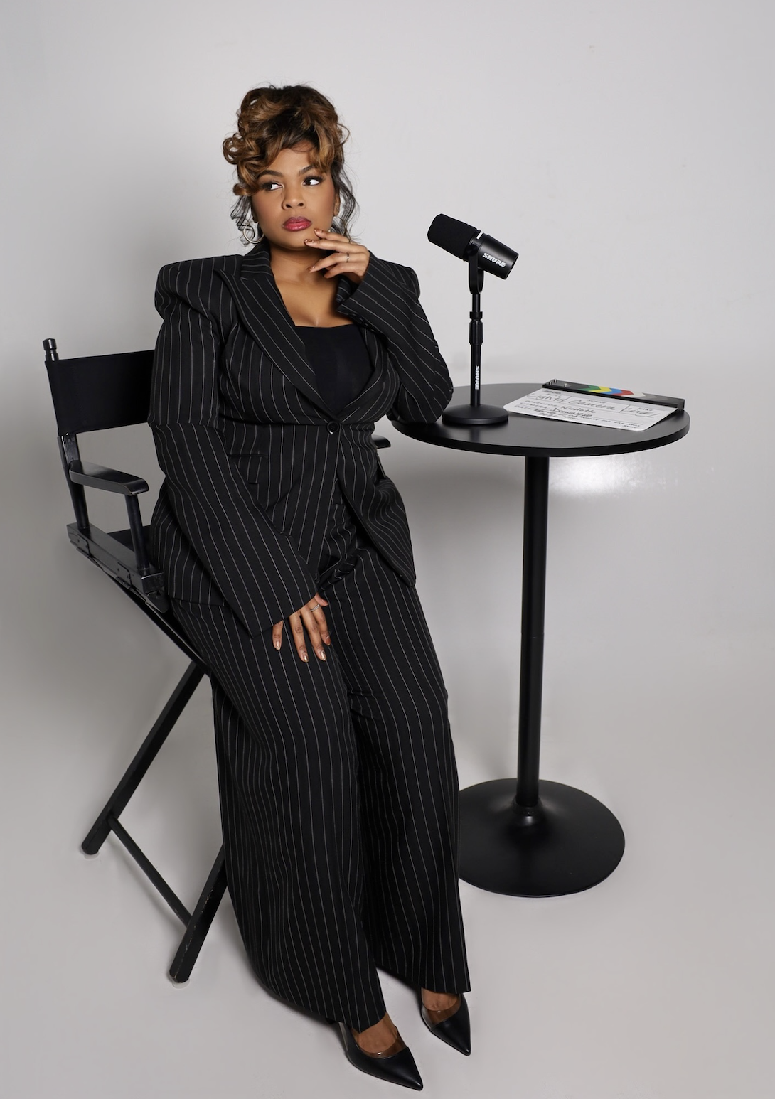 A woman in a black pinstripe suit sitting on a director's chair next to a small round table with a microphone, a script, and some colored markers, against a plain white background.