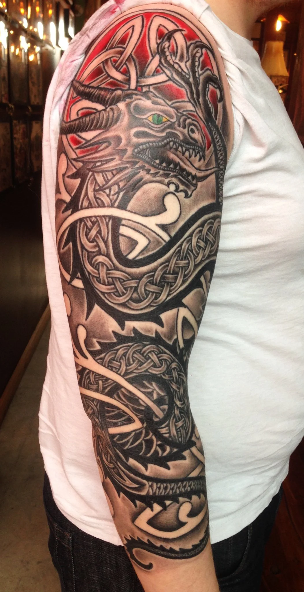 A detailed black and gray sleeve tattoo featuring a dragon with green eye, intertwined with Celtic knot designs, covering the arm from shoulder to wrist.