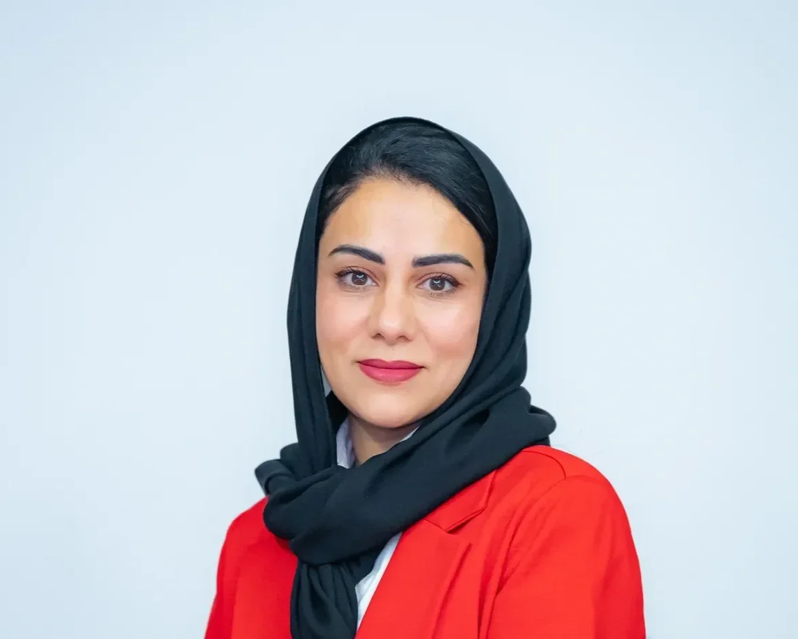 Portrait of a woman wearing a black headscarf and a red blazer against a plain background.