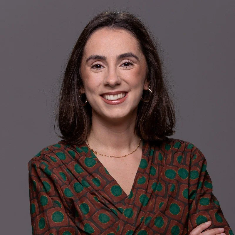 A smiling woman with shoulder-length brown hair, wearing a patterned shirt with green and brown circles and squares, standing against a gray background.