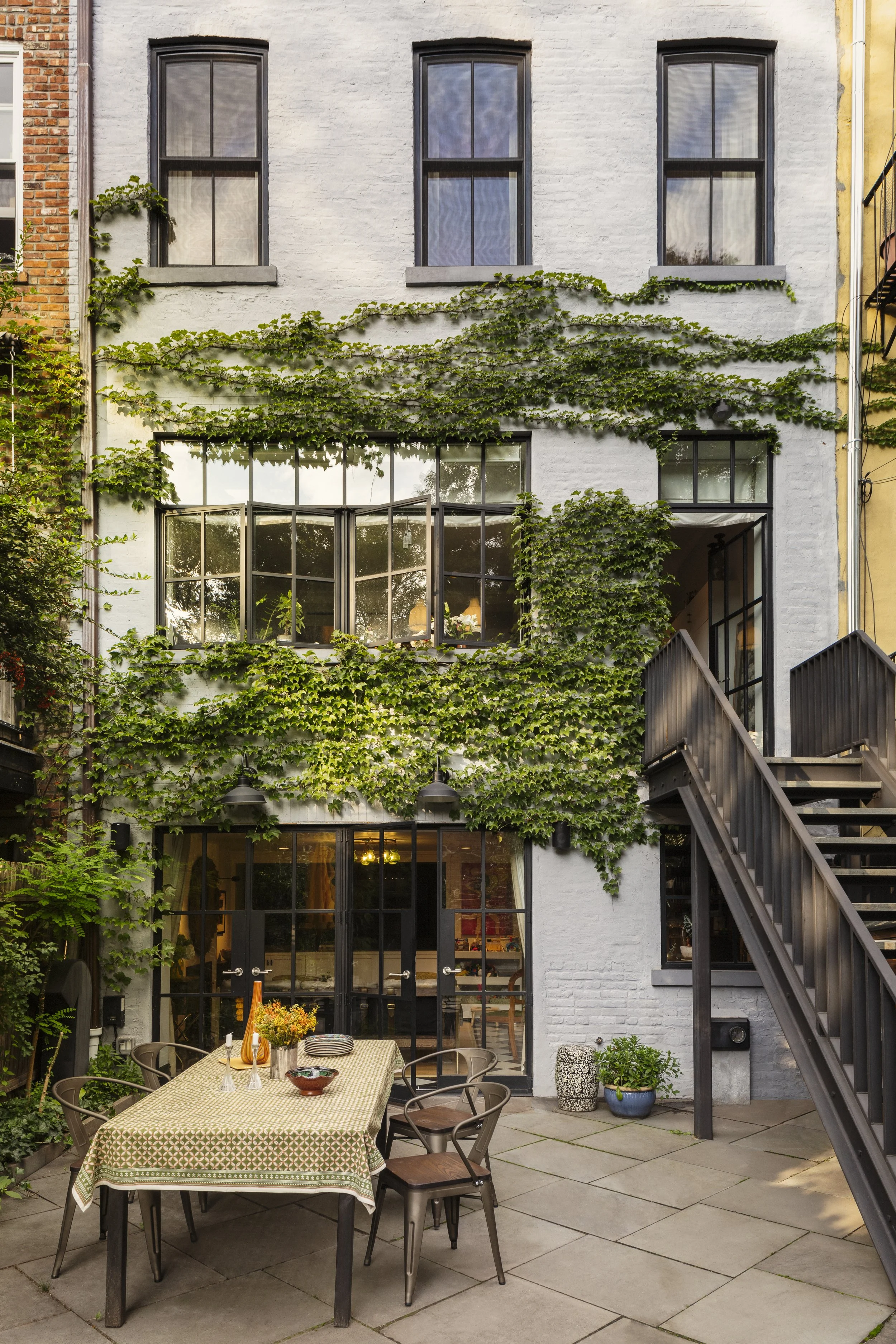 An outdoor patio with a dining table set with plates and a floral centerpiece, surrounded by chairs, in front of a white brick building with black-framed windows and green ivy climbing the walls.