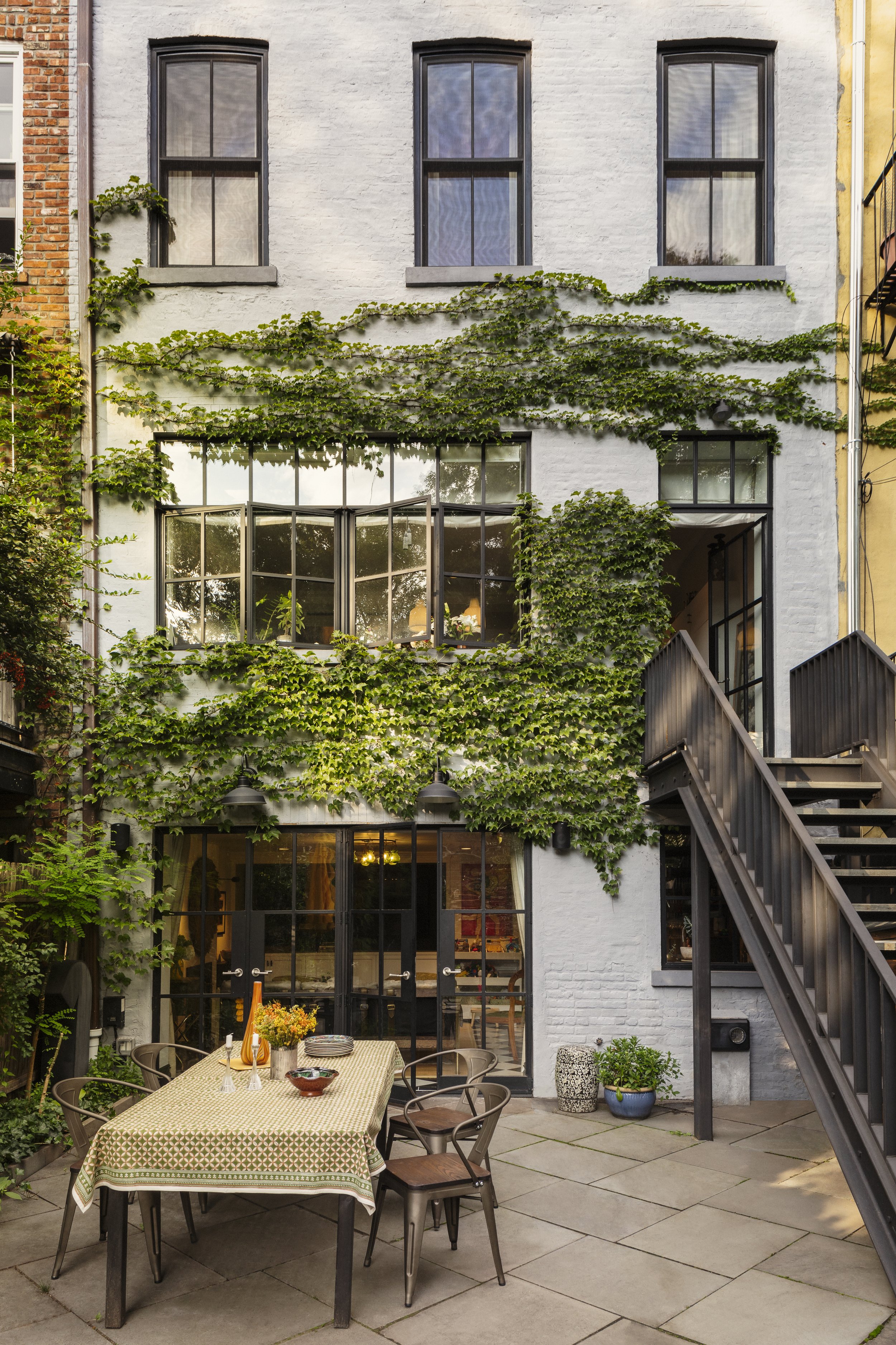 Outdoor patio area with a dining table, chairs, potted plants, and a building with green ivy and large windows.