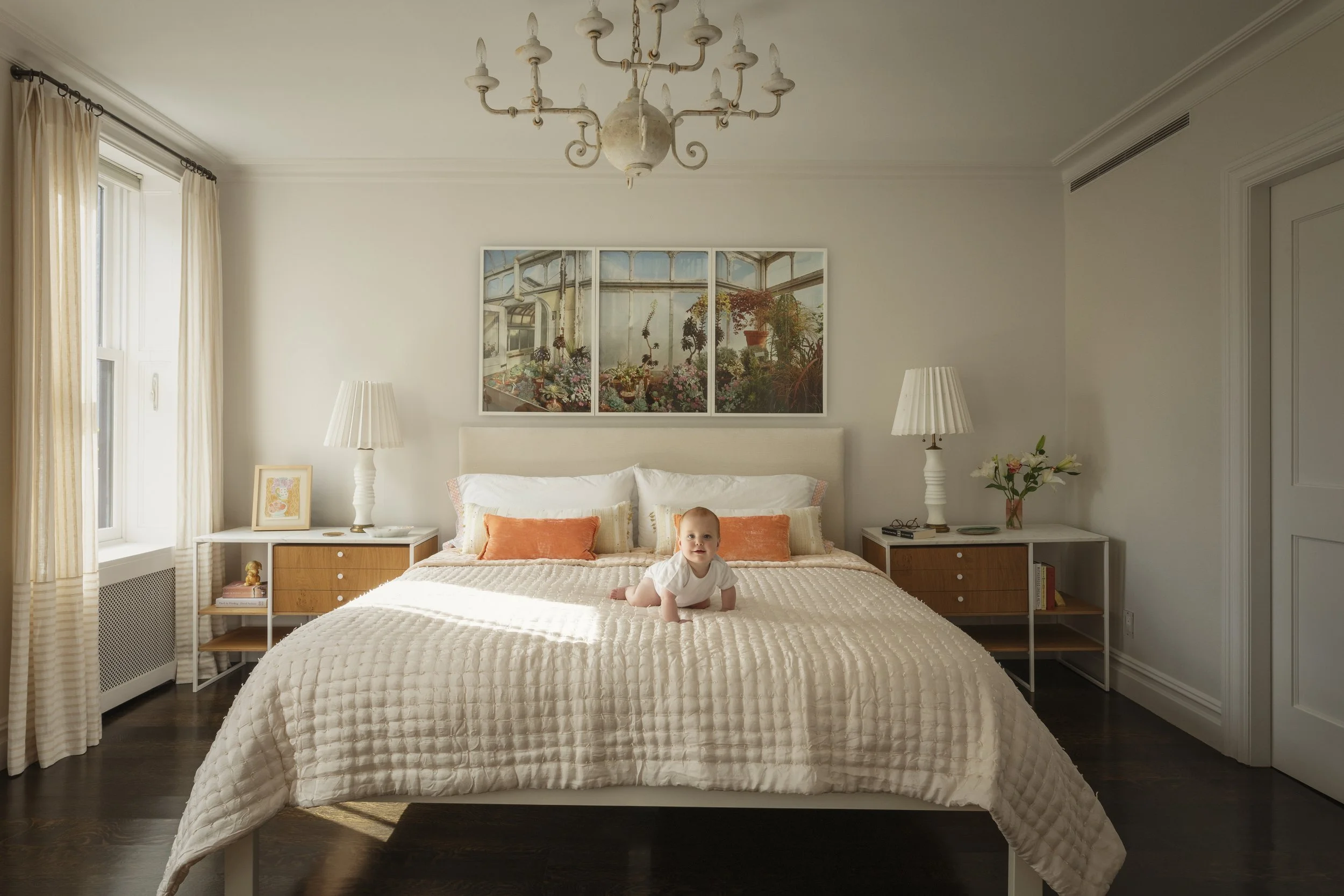 A baby crawling on a large bed in a bright, modern bedroom with white walls, dark wood flooring, and cream curtains. There are two white bedside tables with lamps, a flower vase, and framed artwork above the bed.