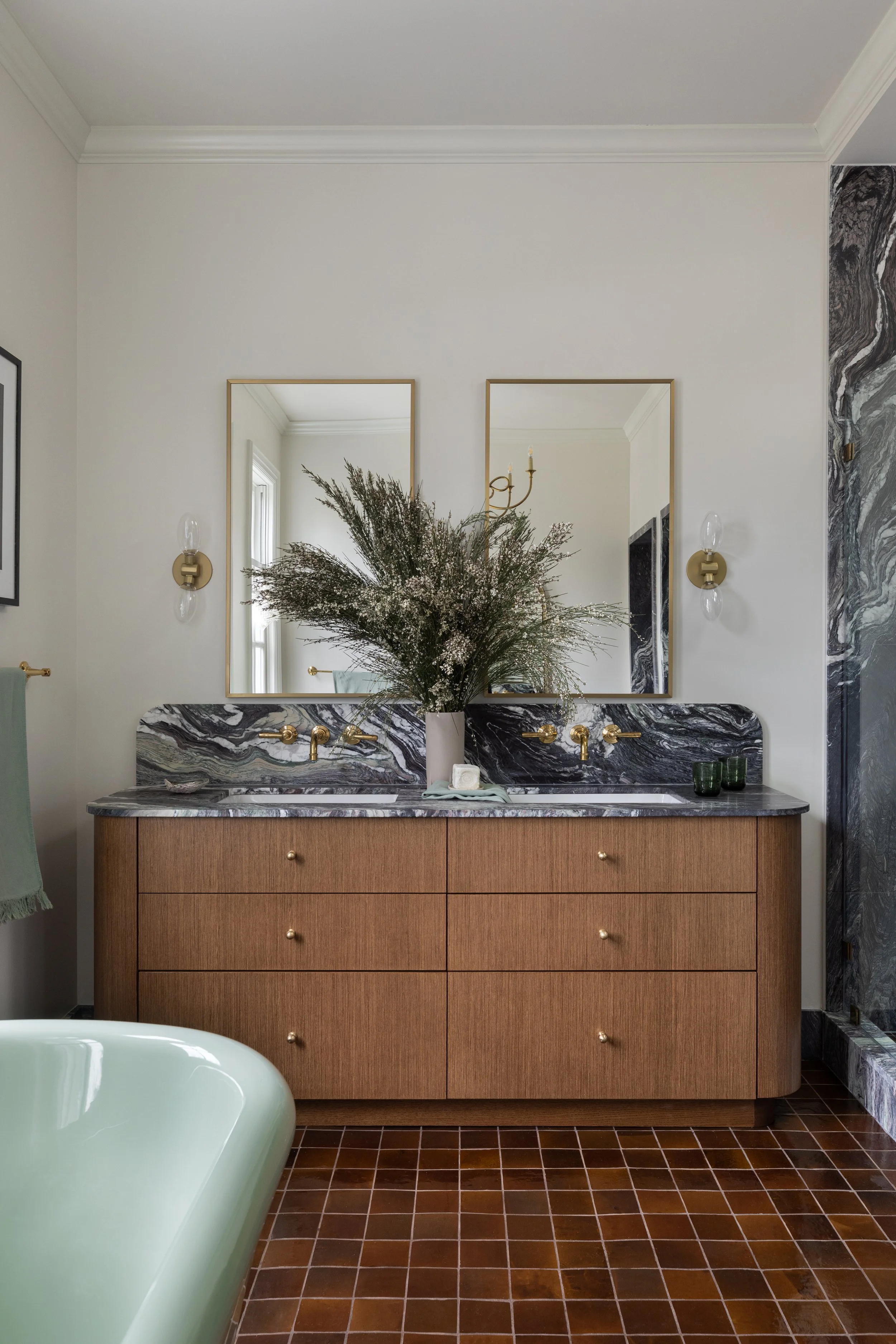 A bathroom vanity with a wooden cabinet, two sinks, and black and white marble countertops, decorated with a large vase of dried flowers, and reflected in two mirrors with gold framing, with a side wall featuring black and white marble, and a brown tiled floor.