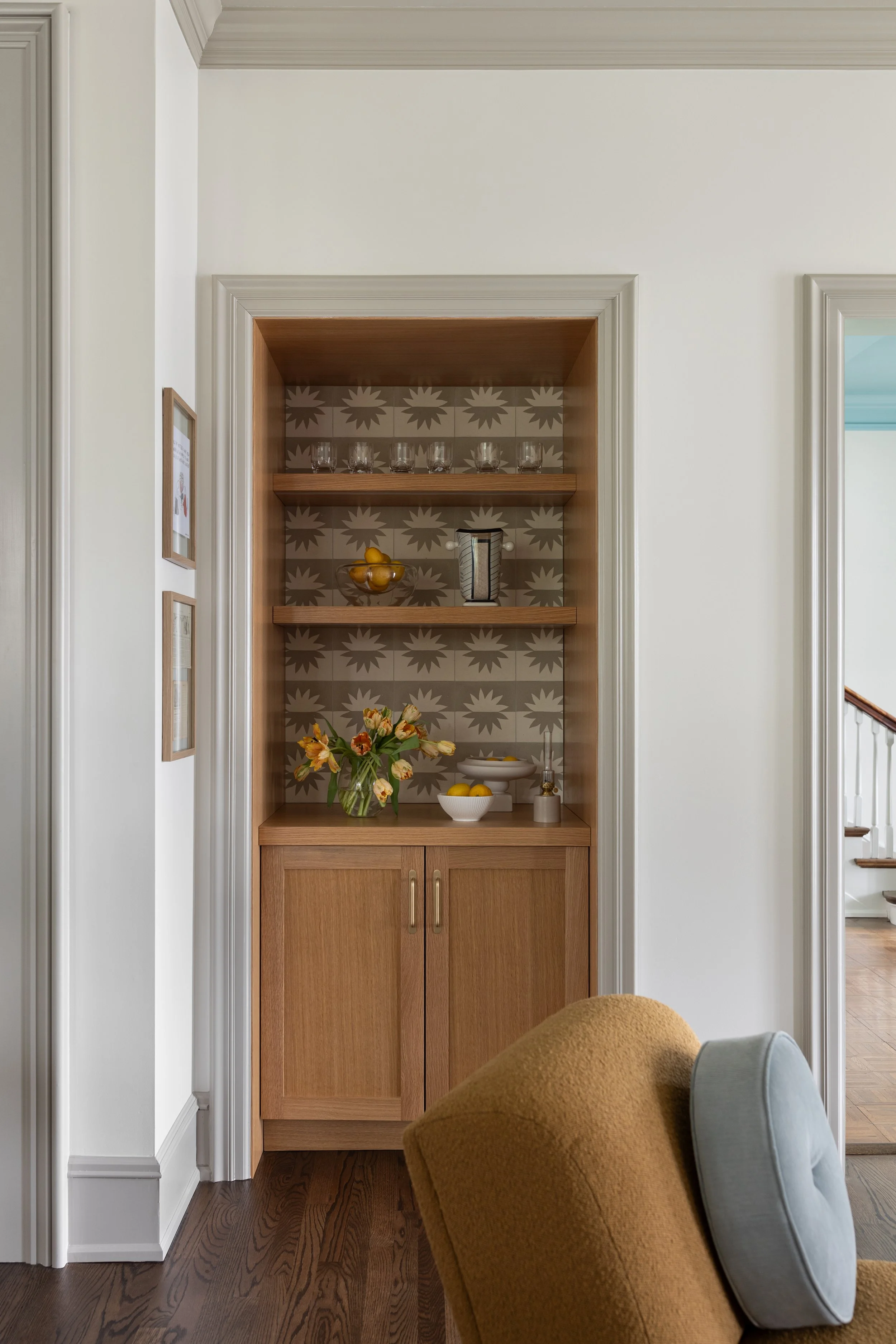 Built-in wooden display cabinet with decorative starburst-patterned backsplash, glassware on top shelf, bowl of lemons, coffee jar, flowers, and bowls on middle shelf, with a closed lower cabinet, in a living room with a chair and framed artwork on the wall.