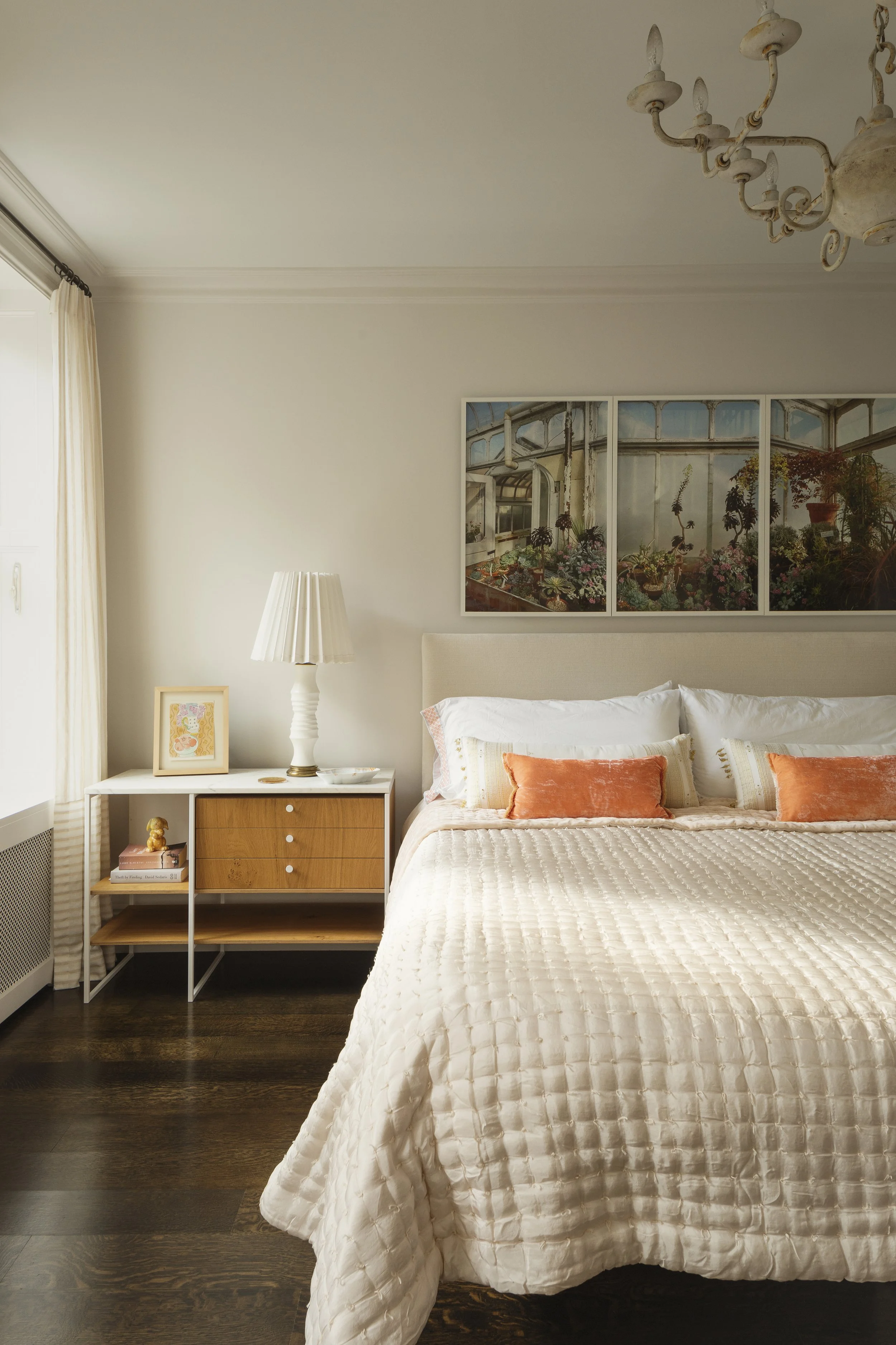 A cozy bedroom with a white bed covered in a textured quilt and decorated with orange pillows. Next to the bed is a white nightstand with a table lamp, framed artwork, and a small decorative object. Above the bed hangs a large framed picture of a greenhouse interior filled with plants. The room has dark wood flooring and cream-colored walls, with a window covered by light curtains.
