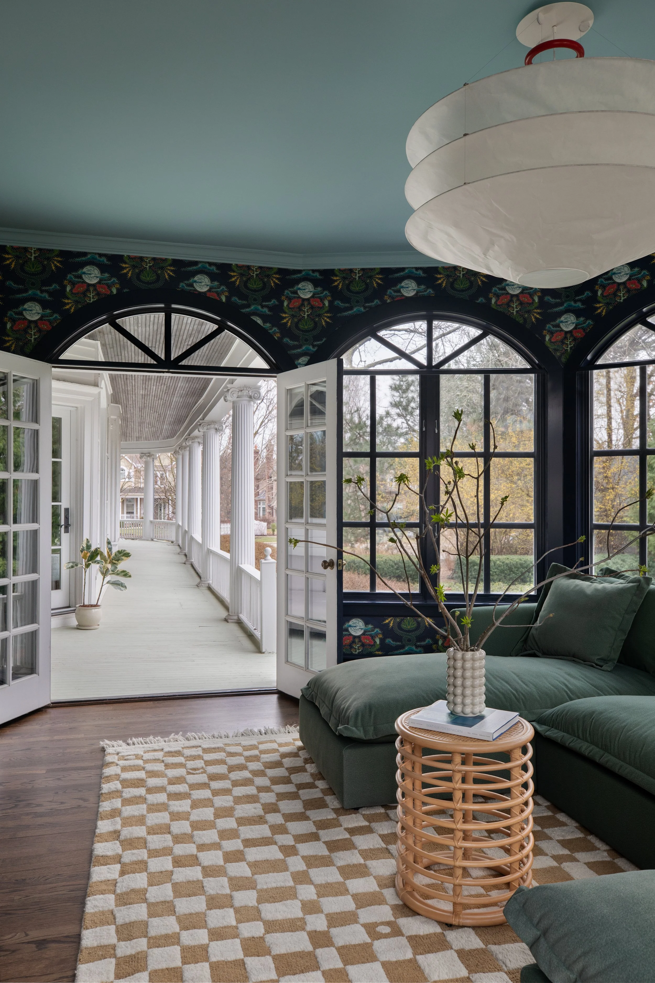 Living room with large windows, showing a porch with white columns outside. The room features a green sofa, a rattan side table with a vase and books, a checkered rug, and a ceiling lamp.