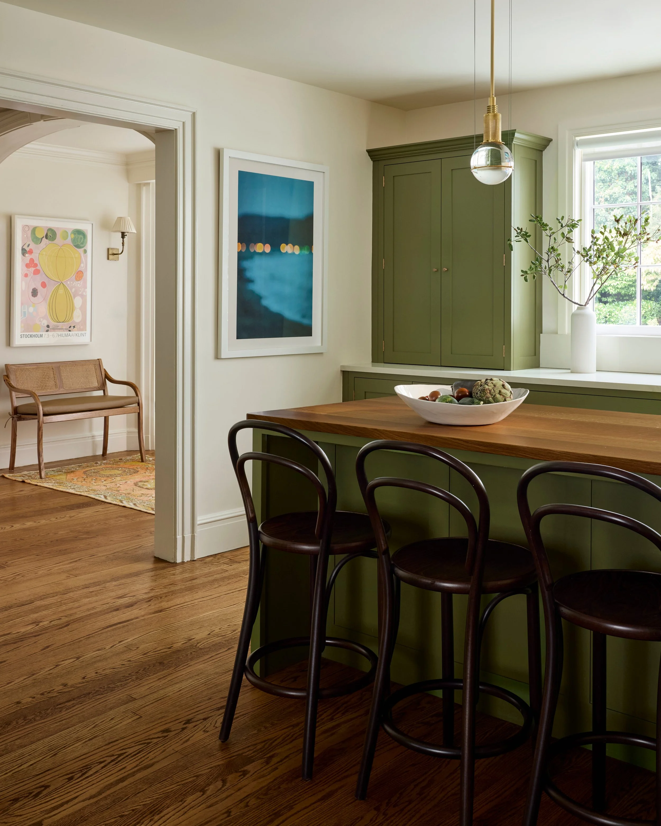 A kitchen with a green island, black bar stools, wooden flooring, and wall art. A window with a vase and plant on the sill. Adjacent room has a wooden bench and framed artwork.