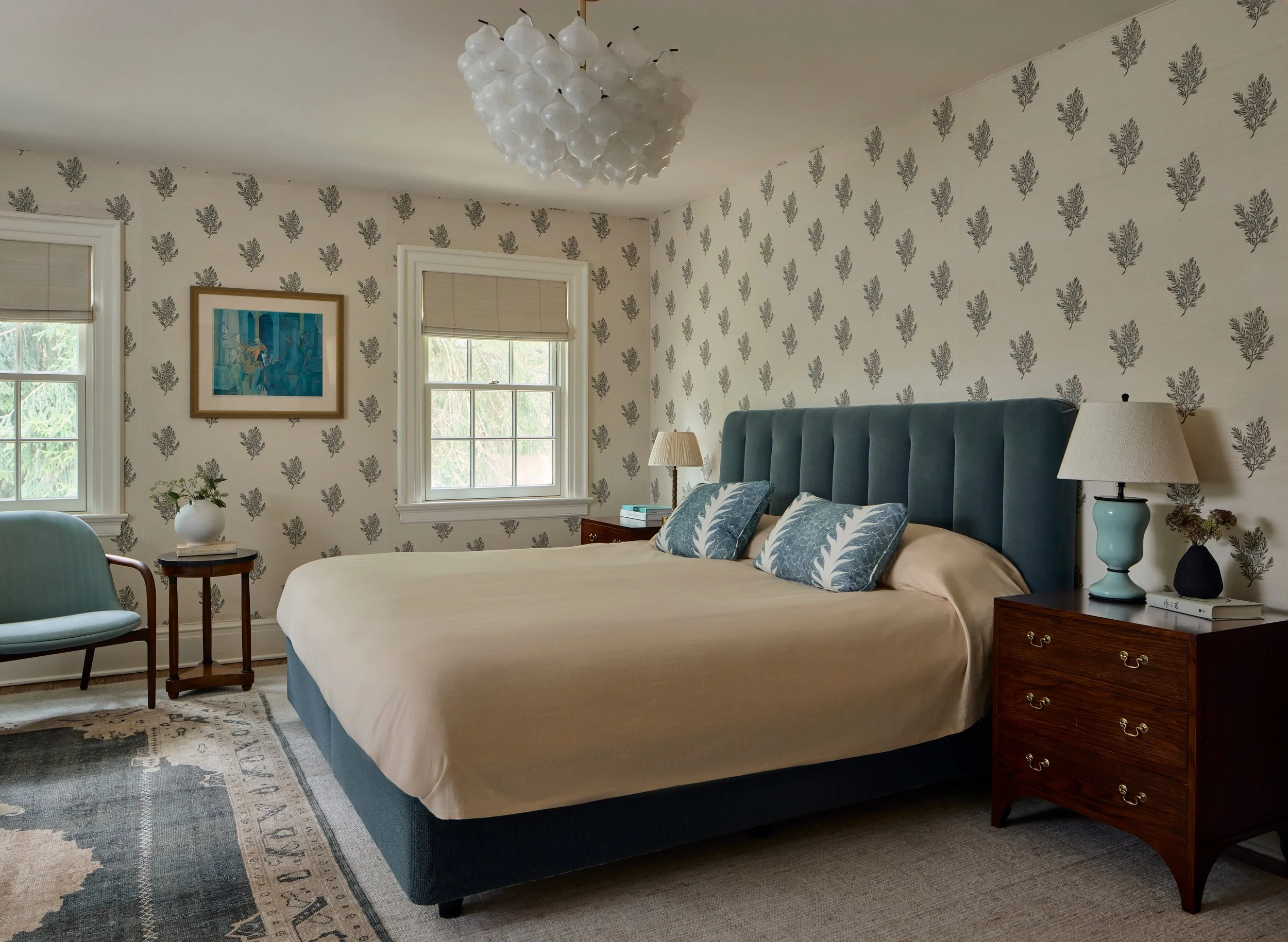 A cozy bedroom featuring a large bed with a teal velvet headboard, beige bedding, and decorative pillows. The room has patterned wallpaper with gray leaves, two windows with white framing, and matching beige blinds. There are two wooden nightstands with lamps, a small side table with a vase of flowers, a light blue armchair, and a rug with a classic design. A modern ceiling light fixture hangs in the room.