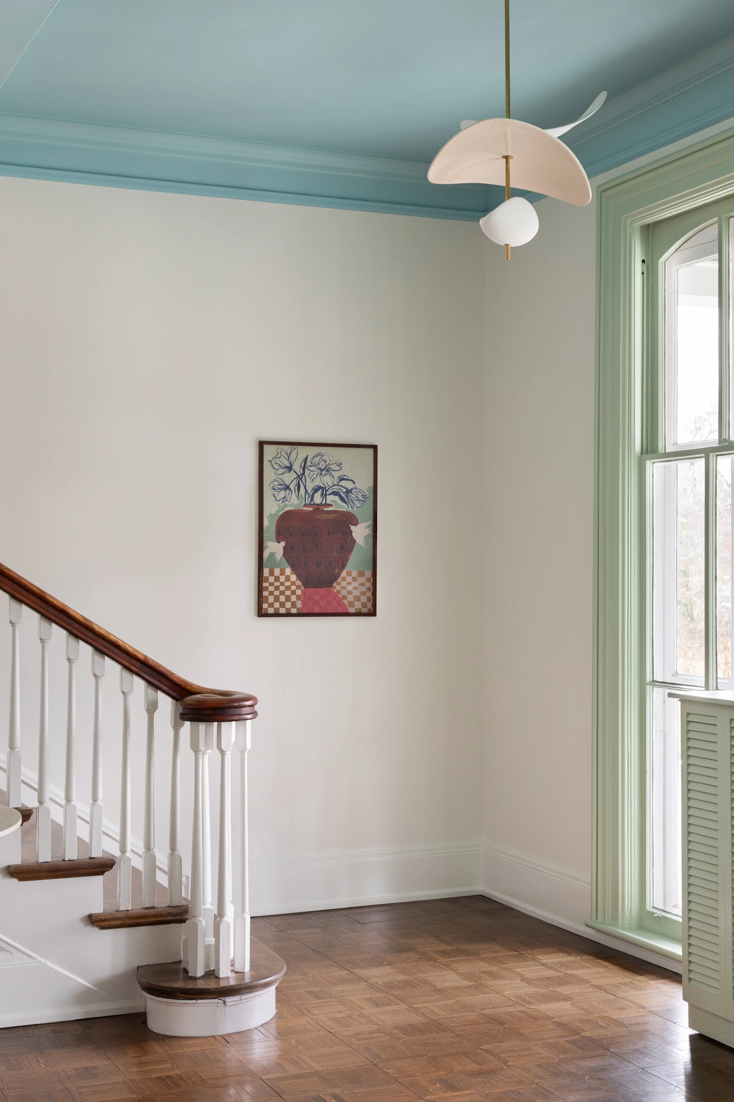 Interior of a room with a staircase, a large window with green trim, a hanging modern light fixture, a framed artwork of a flower vase, and hardwood floors.