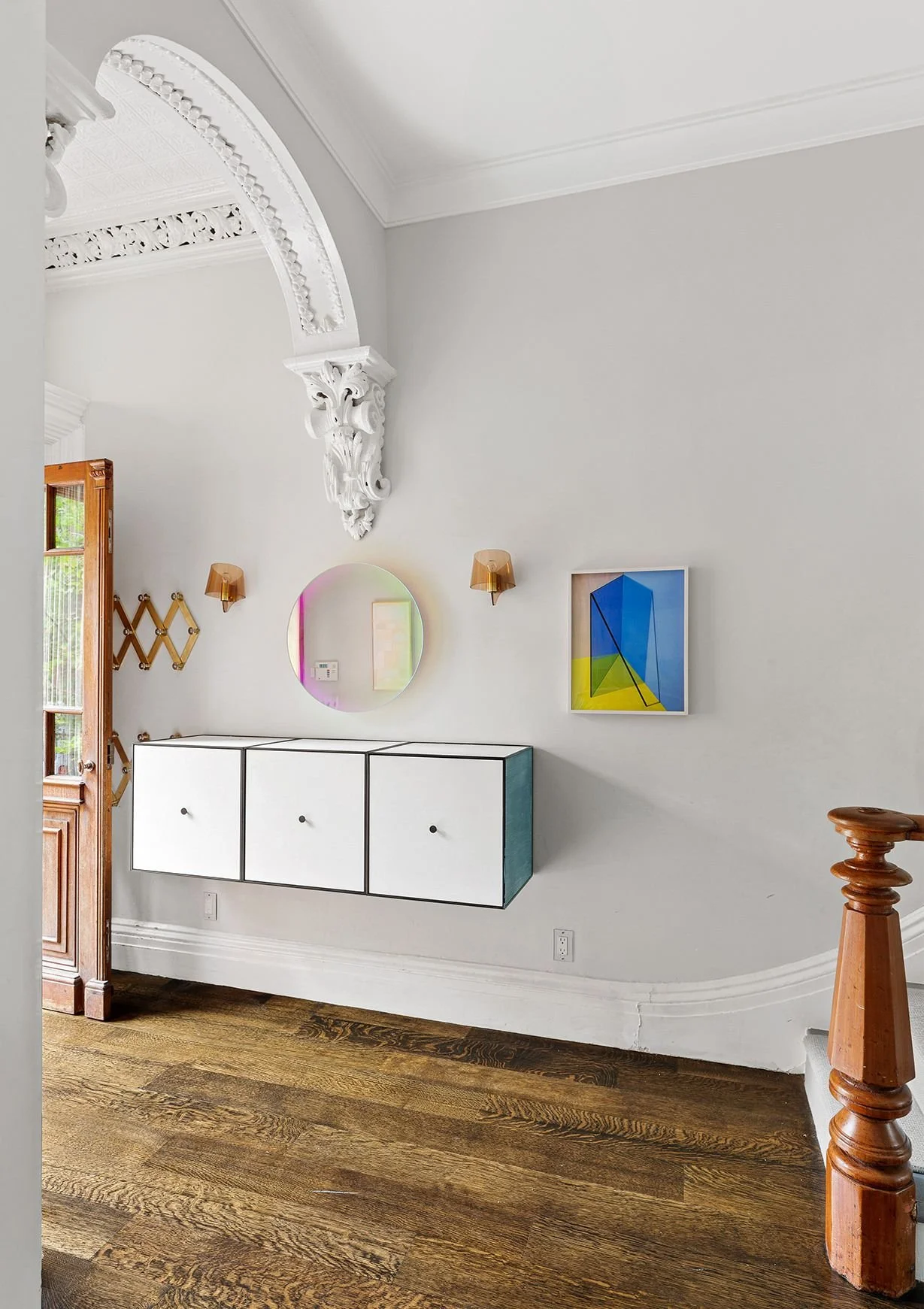Interior of a house entryway with a wooden door, white walls, a floating black-and-white cabinet, decorative mirror, colorful abstract artwork, wall sconces, and wooden staircase post.