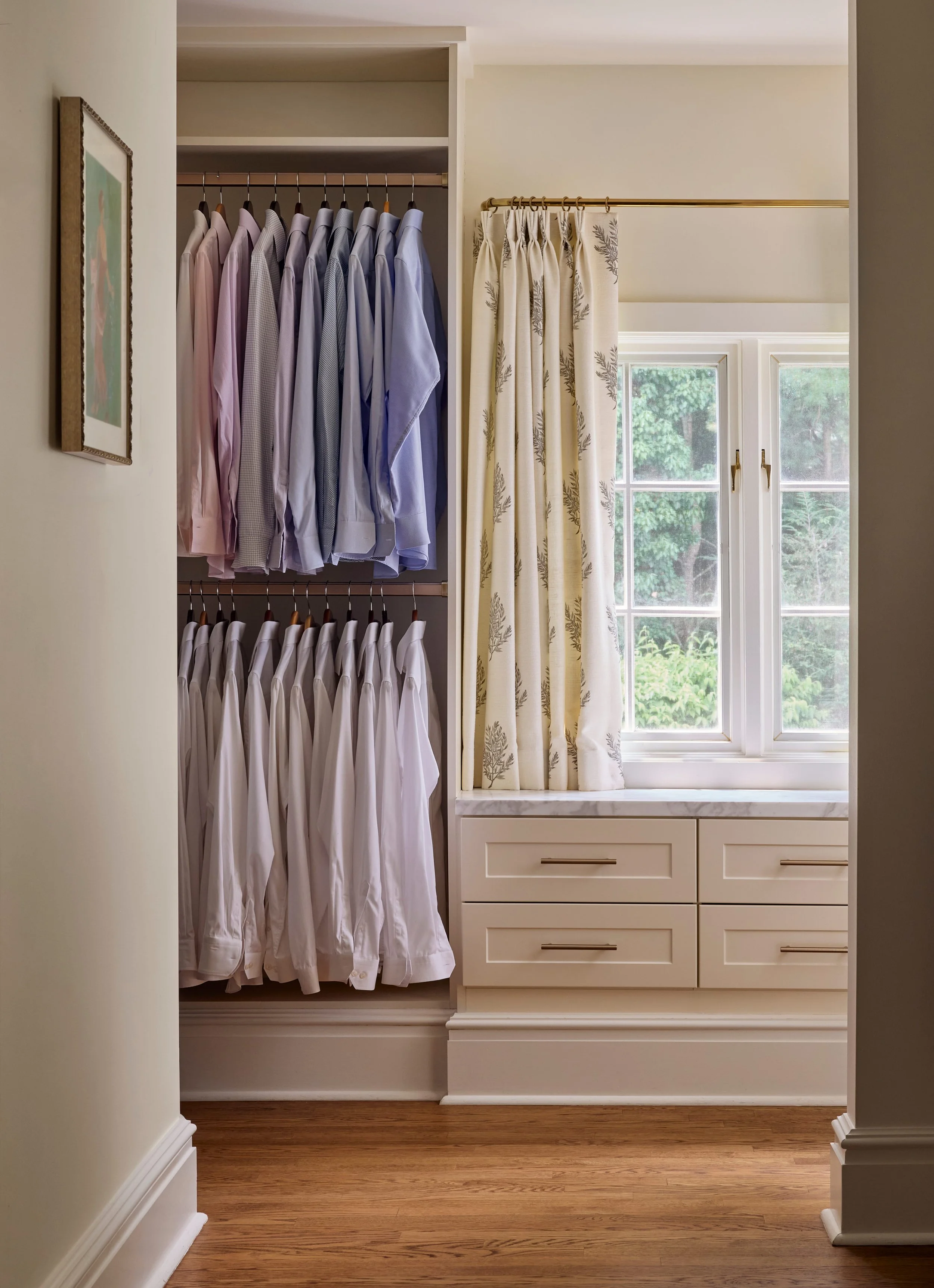 Closet with two racks of shirts, a window with cream curtains, and built-in drawers underneath the window.