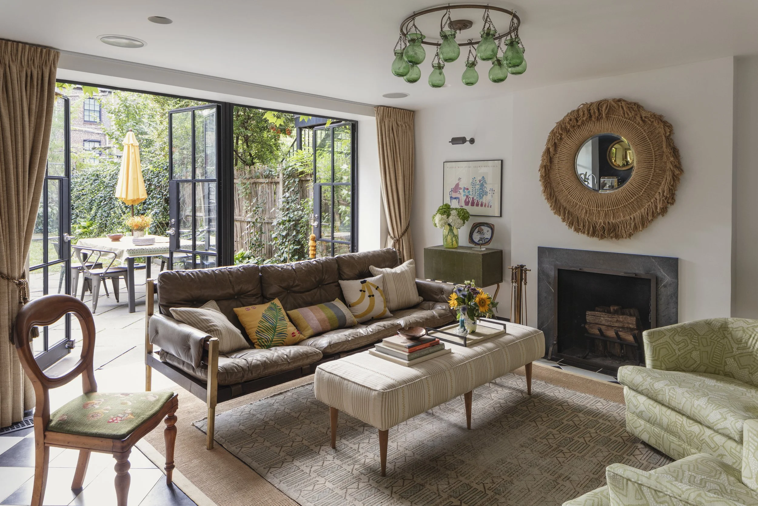 Living room with a brown leather sofa, a striped ottoman with books and a vase of flowers, a green armchair, a fireplace with a woven wall hanging mirror, and large glass doors opening to an outdoor patio with a table, chairs, and yellow umbrella.