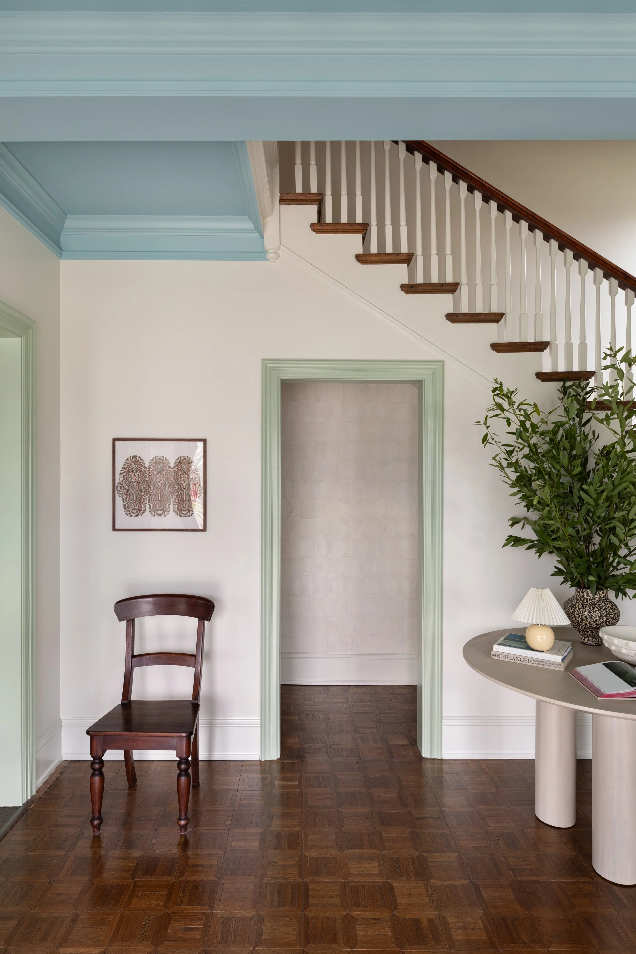 Interior of a home featuring a staircase with wooden steps and white spindles, a small wooden chair, a side table with a large plant, books, and decorative items, and artwork on the wall.