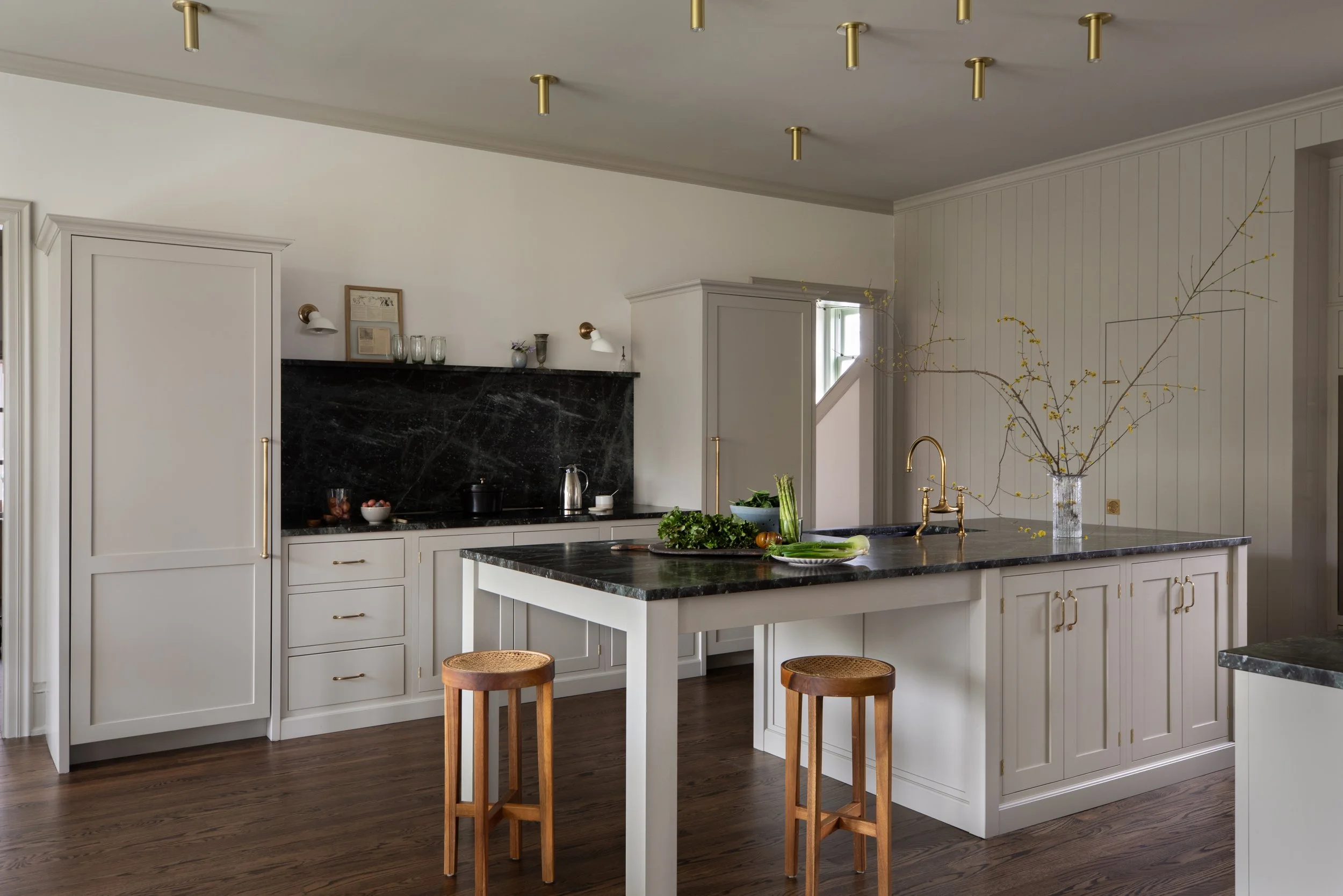 Modern kitchen with white cabinets, black marble countertops, and gold fixtures. An island with two wooden stools has a vase with branches and yellow flowers, along with fresh vegetables. The room has wood flooring and a window letting in natural light.