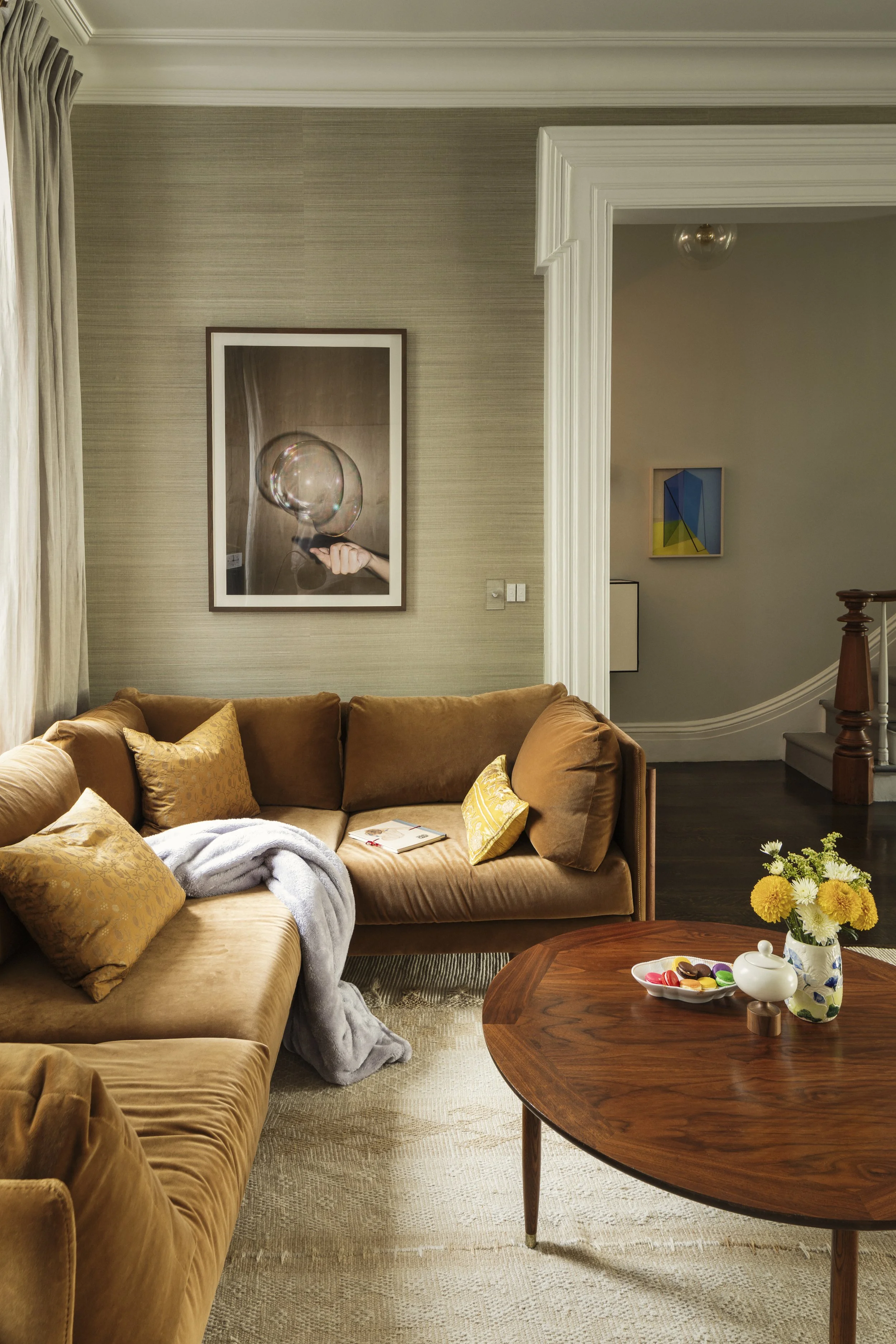 Living room with a brown velvet sectional sofa family room with decorative pillows, gray blanket, a wooden coffee table with a flower vase, a plate of colorful macarons, and artwork on the walls.