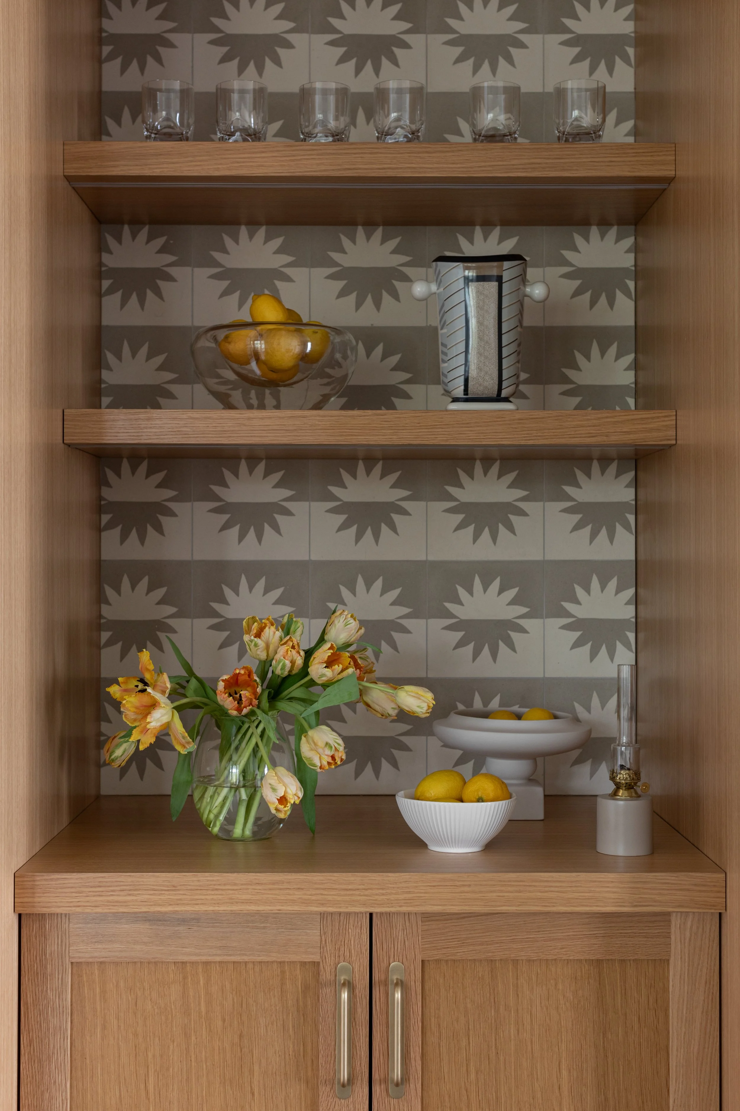 A wooden kitchen cabinet with open shelves decorated with glassware, lemons in a bowl, a floral vase with tulips, and a vintage oil lamp, with a patterned tile background.