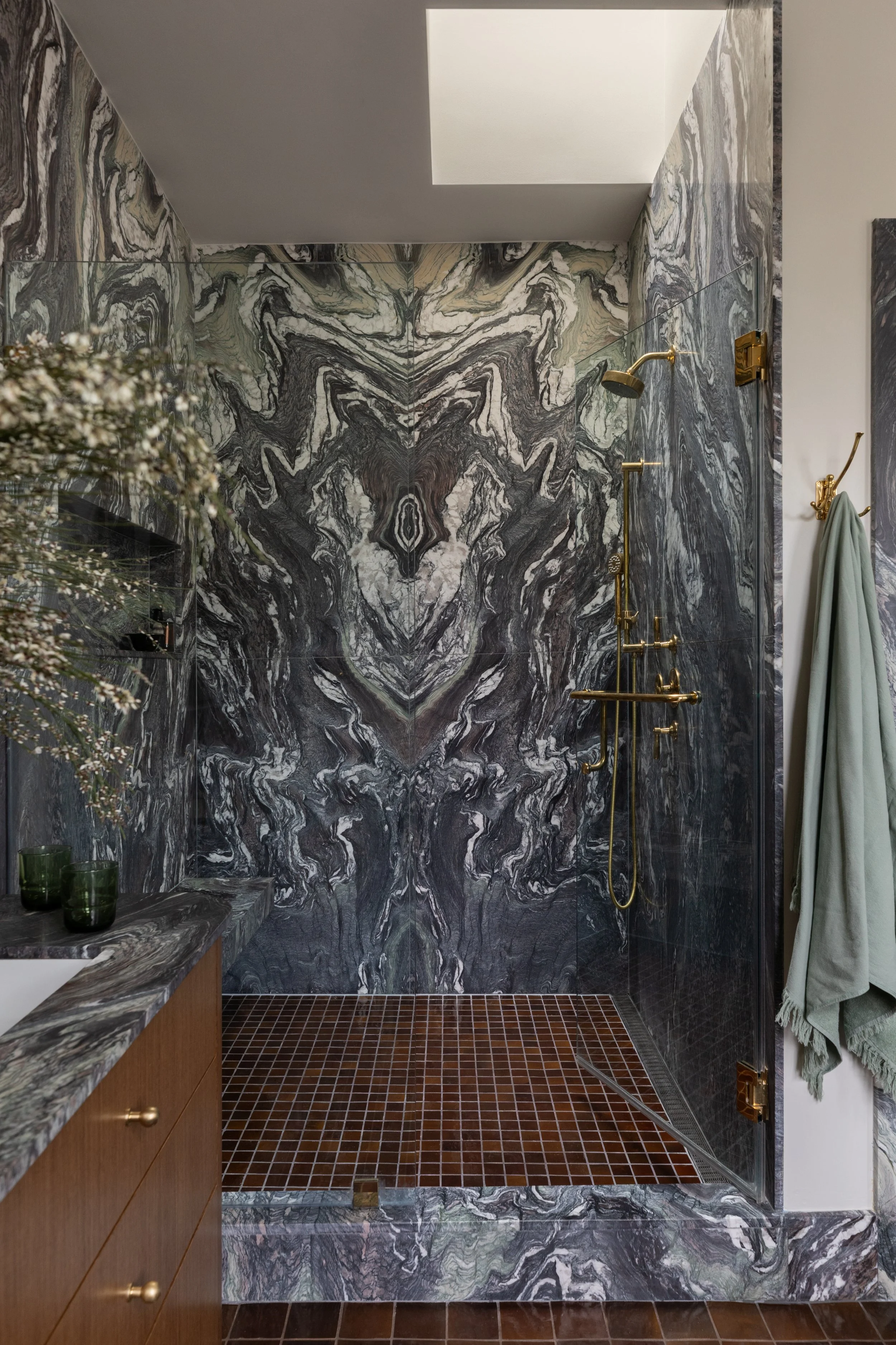 Bathroom shower with black and white marble walls, gold fixtures, a glass door, a skylight, and a dark red tiled floor.