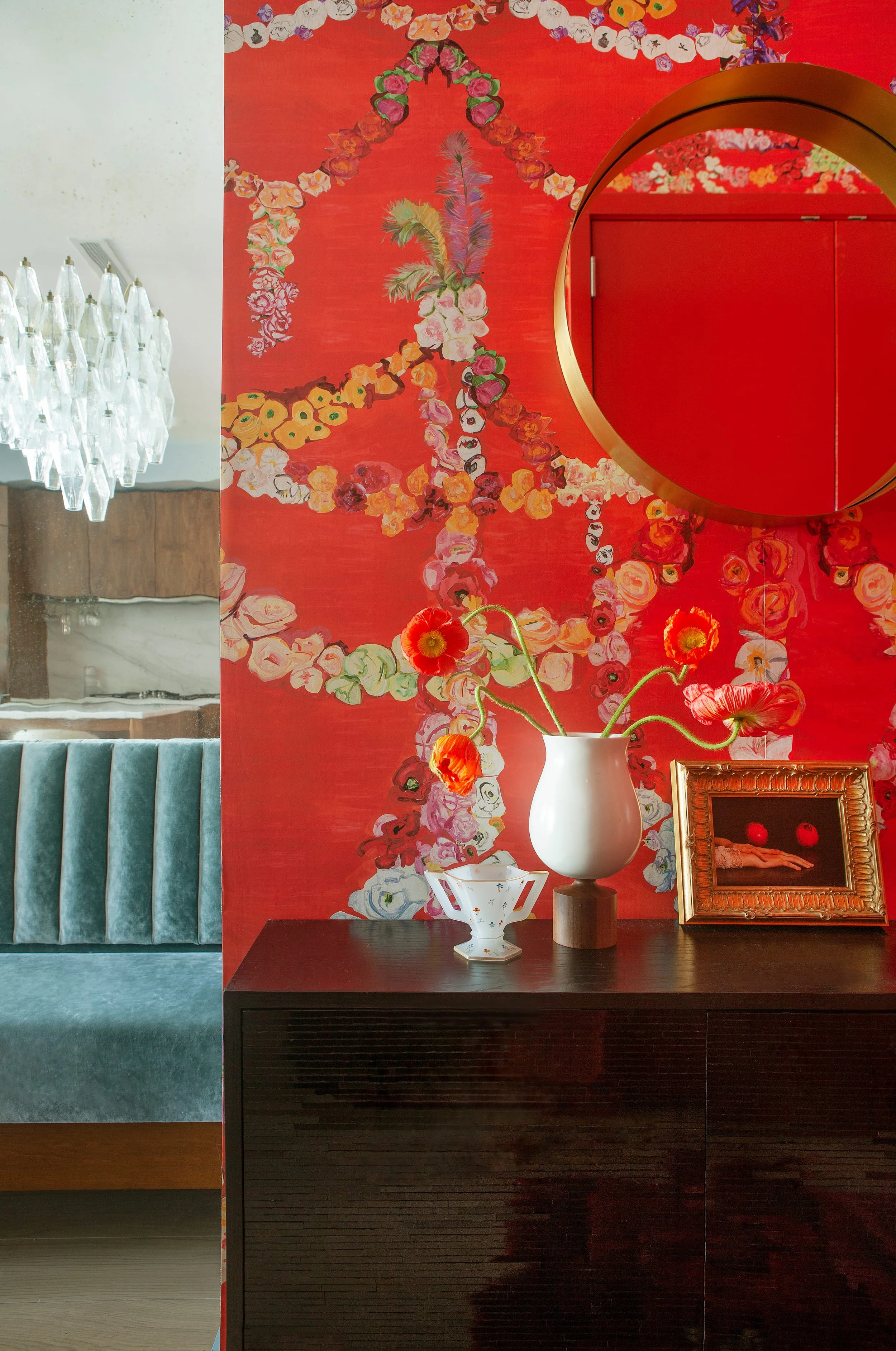 Interior decor with a dark wooden sideboard holding a white vase with red and orange flowers, a small decorative cup, and a framed art piece. Behind it, a circular mirror and a vibrant red floral wall with pink, white, and purple flowers.