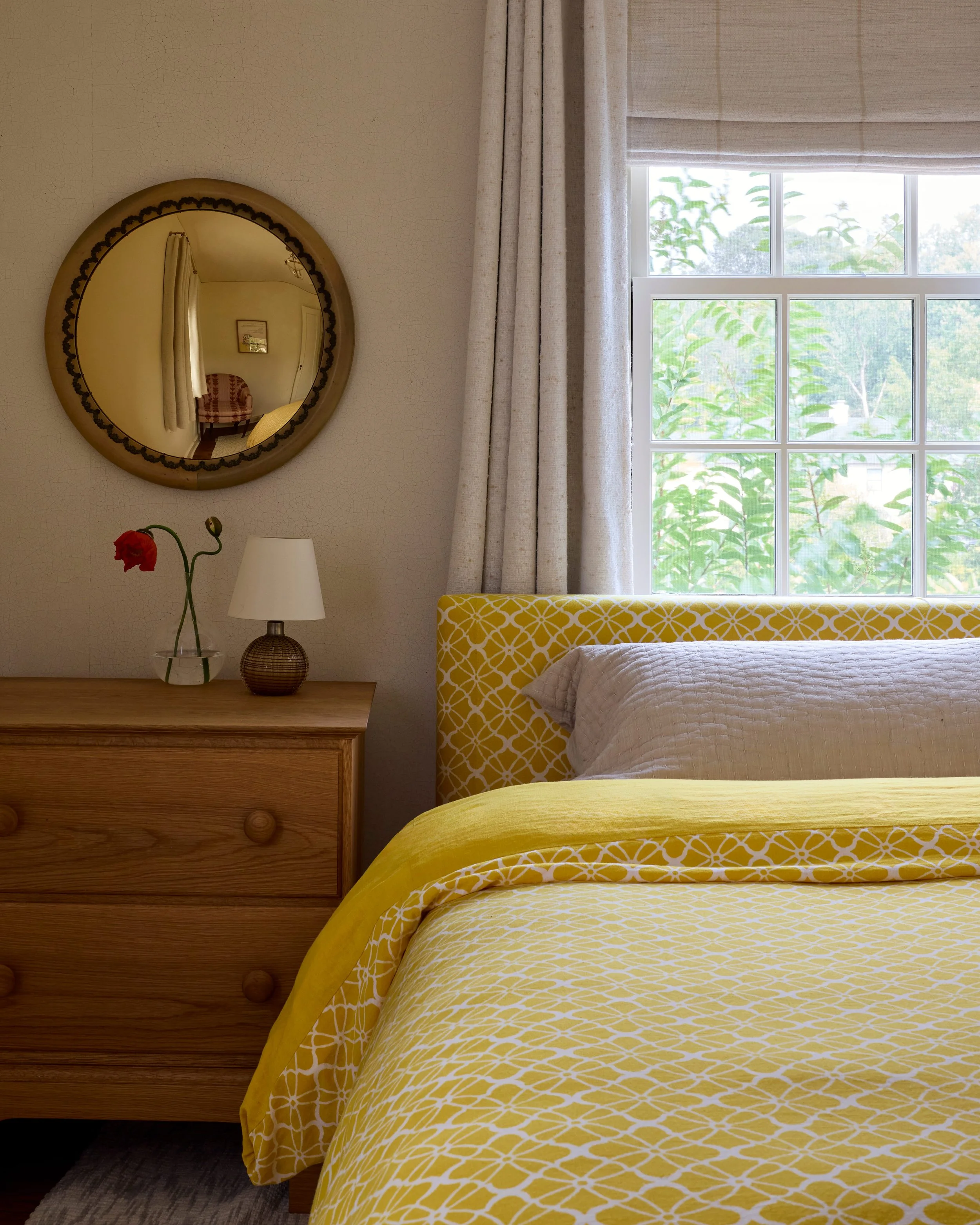 A cozy bedroom with a window showing green trees outside, a yellow patterned bed, white pillow, beige curtains, a wooden dresser with a vase and lamp on it, and a circular mirror reflecting part of the room.