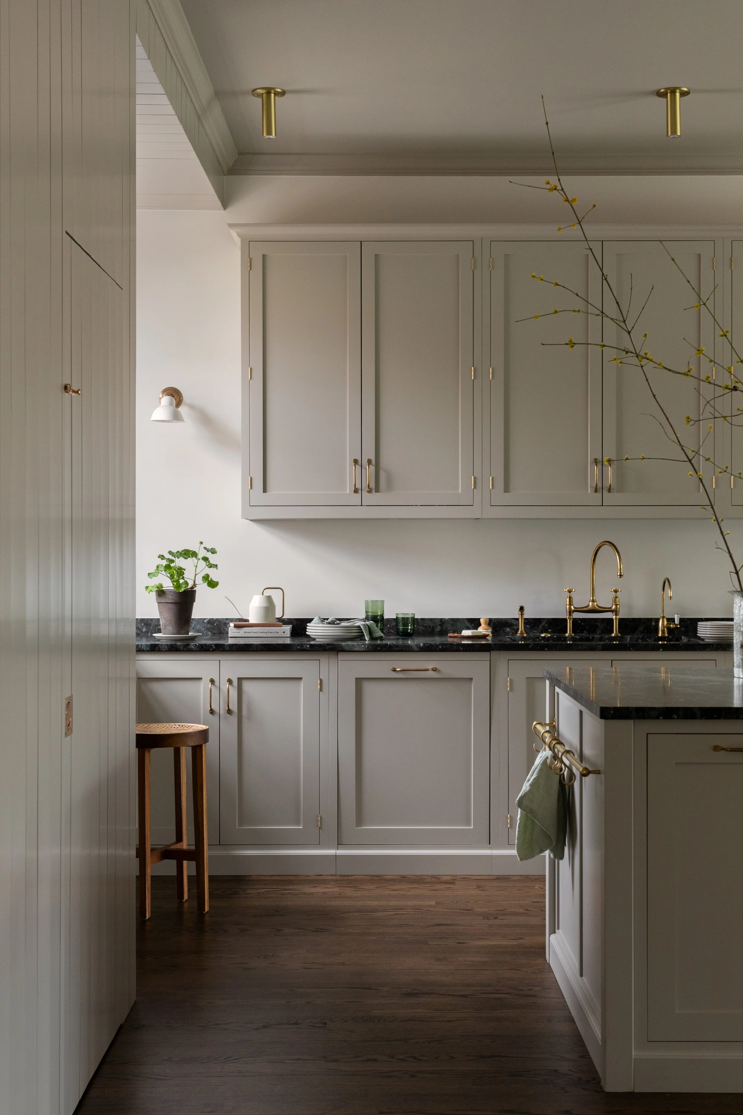 Minimalist kitchen with white cabinetry, black marble countertops, and gold hardware and fixtures, featuring a small potted plant and a vase with branches with yellow flowers.