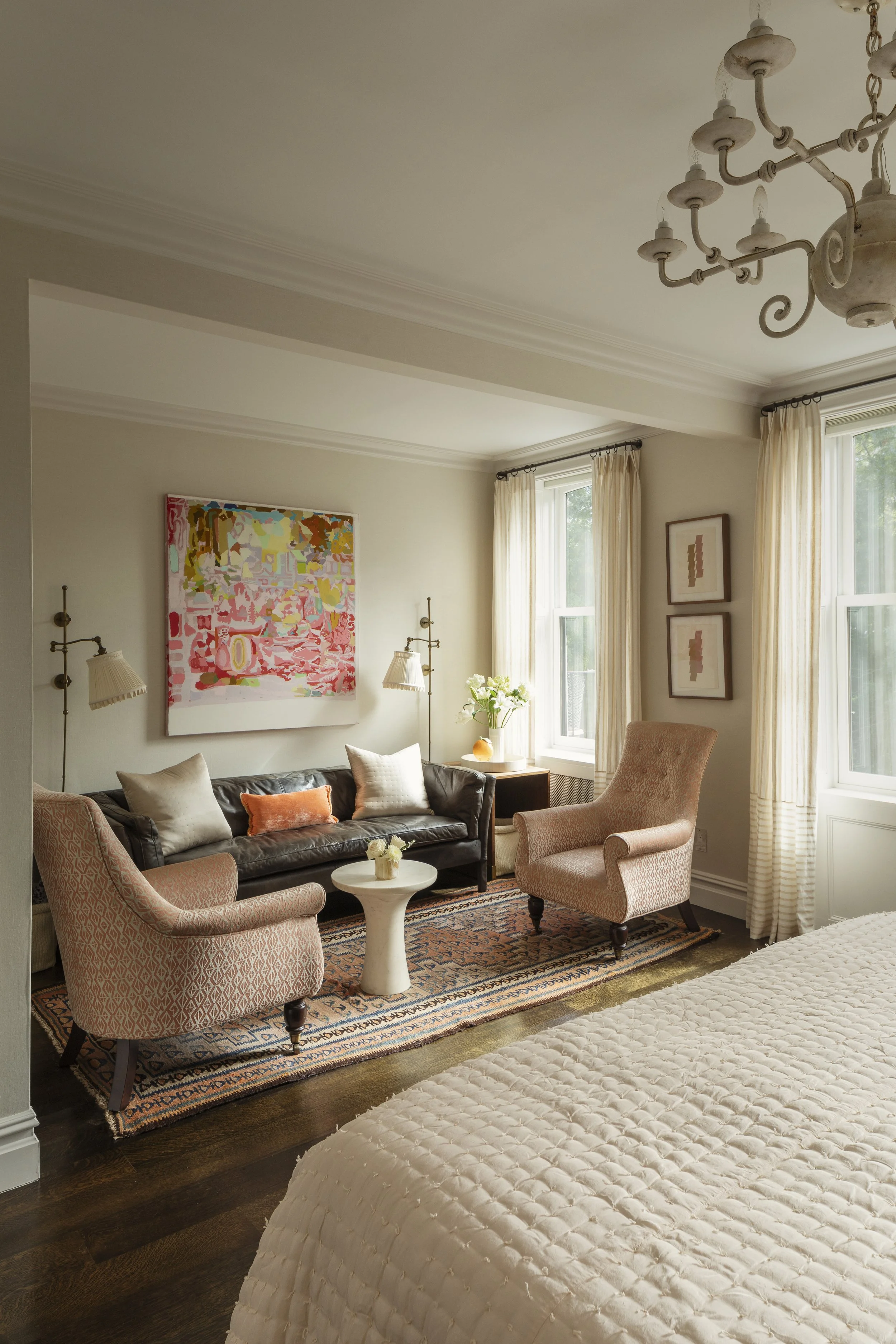 Living room with pink upholstered armchairs, a black leather sofa, a white round coffee table, a colorful rug, artwork on the walls, and two large windows with cream curtains.