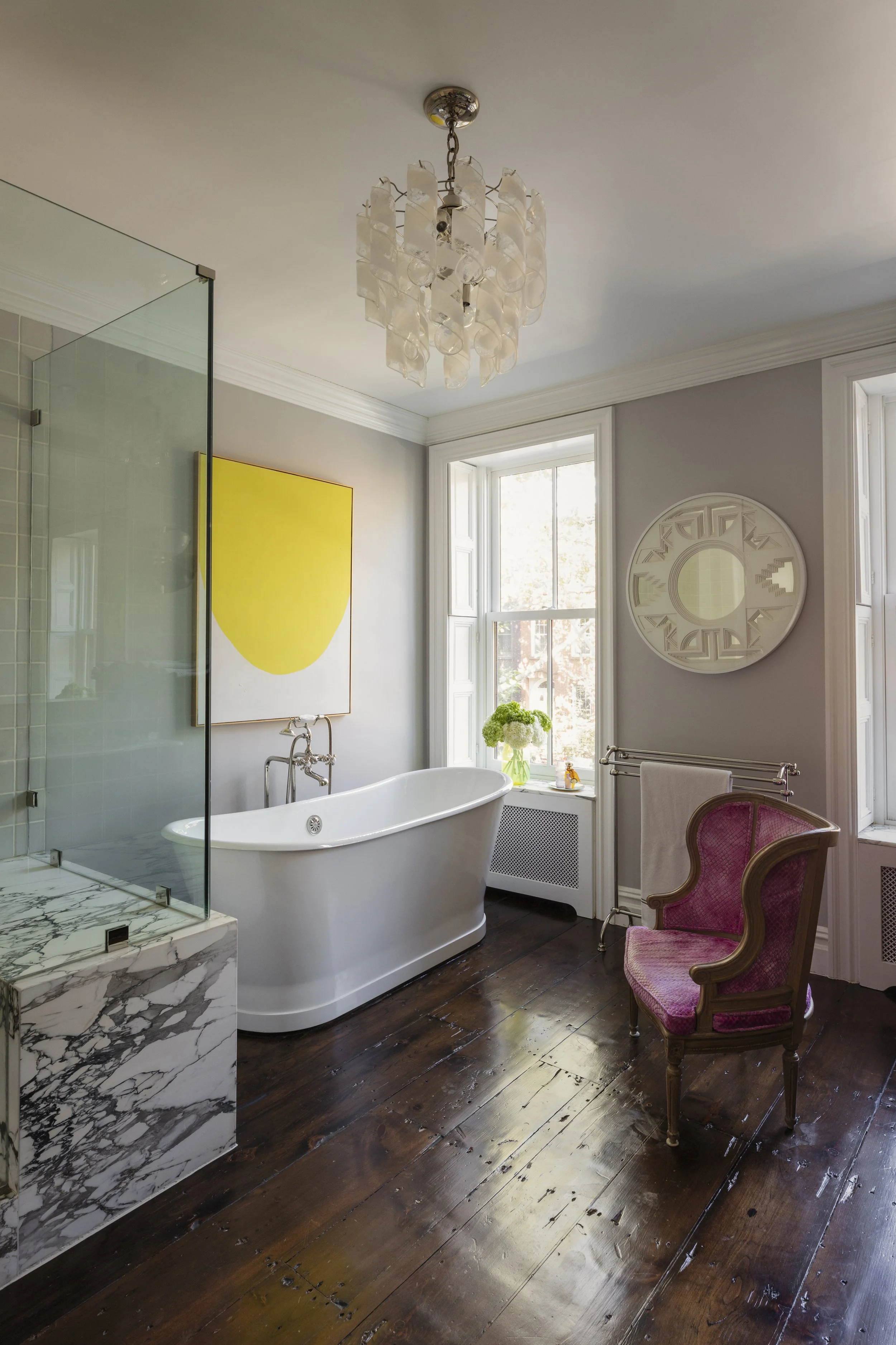 Bright bathroom with a freestanding bathtub, a glass-enclosed shower with marble base, dark wooden floor, large windows, a pink upholstered vintage chair, a vase with green hydrangeas, and modern art on the wall.