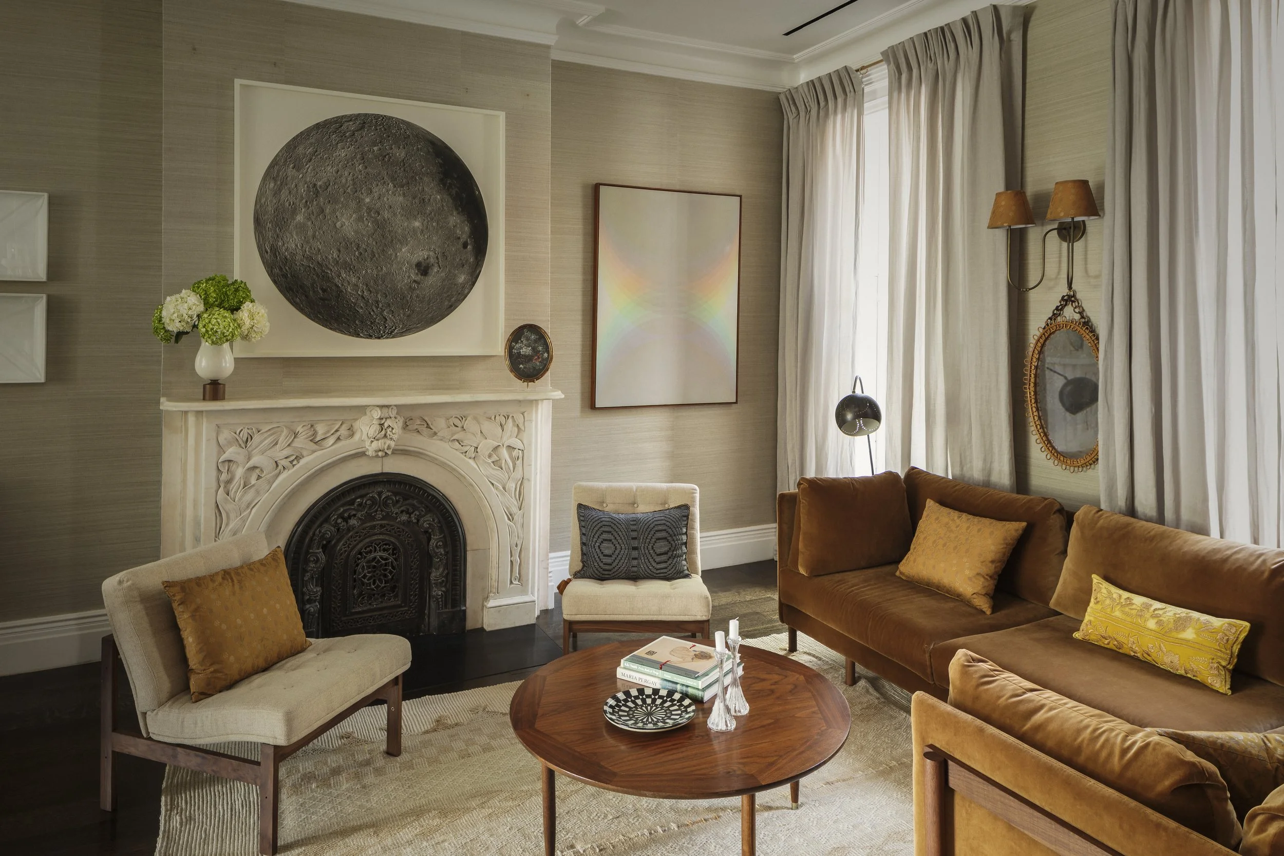Living room with beige walls, white curtains, a brown velvet sofa, cream and gray chairs, a circular wooden coffee table, wall art, a flower vase with white and green flowers, a fireplace, and decorative wall lamps.