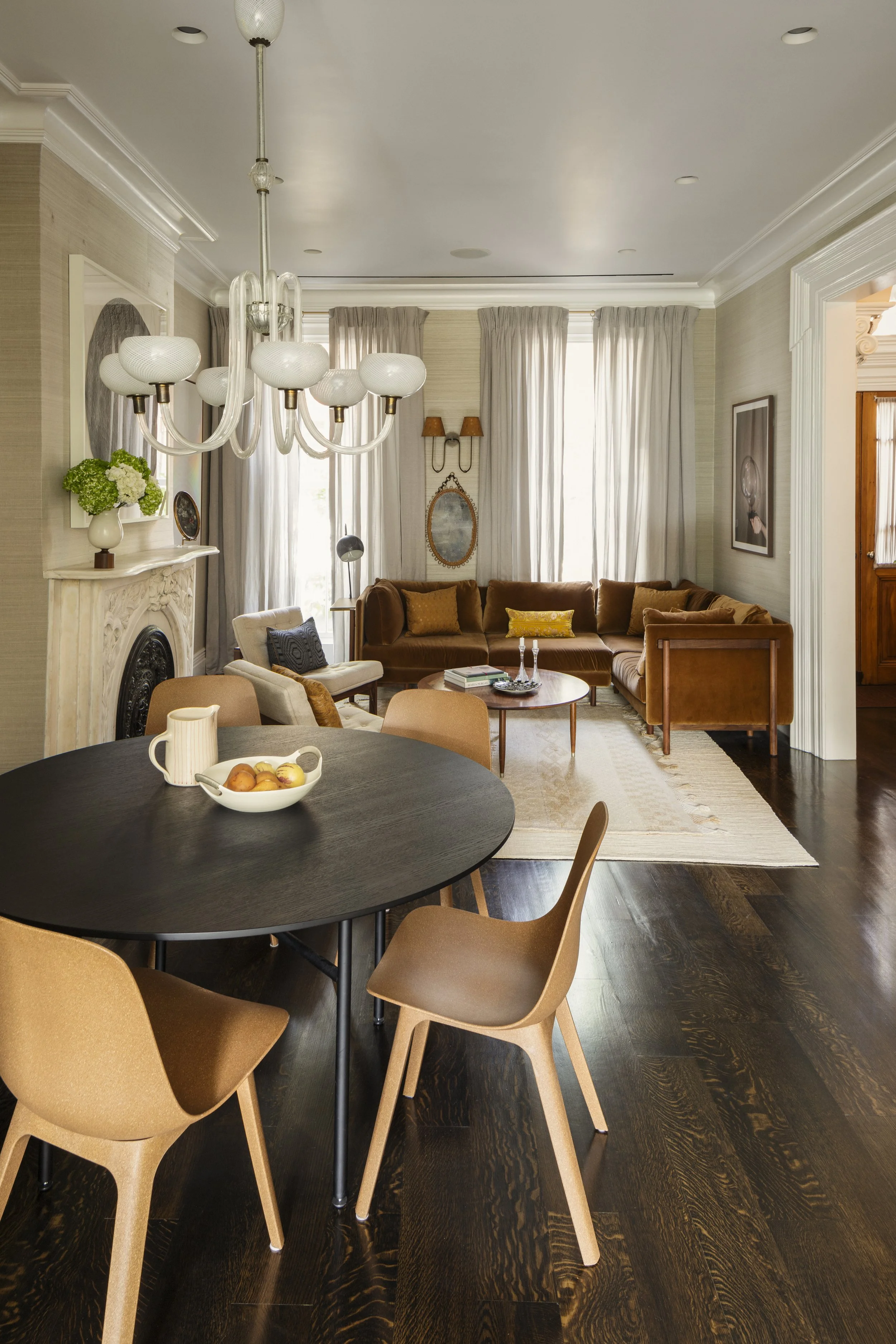 Living room with brown sectional sofa, cream armchair, white fireplace, dark wood floor, black round dining table with beige chairs, chandelier, curtains, and artwork.