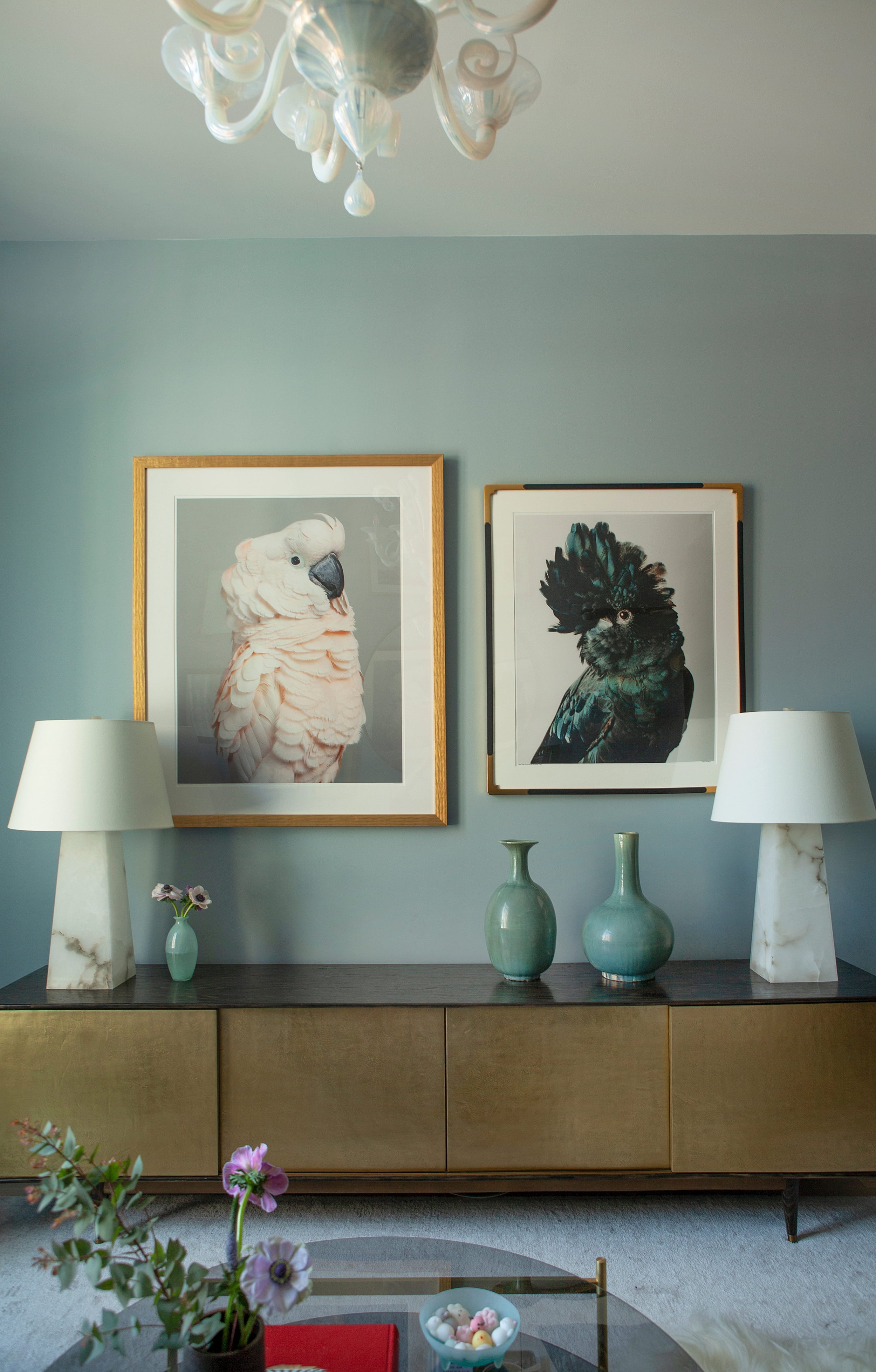 Living room wall with two framed bird photographs, matching white lamps on a wooden sideboard, vases, small flowers, and a glimpse of a coffee table with decorative items.