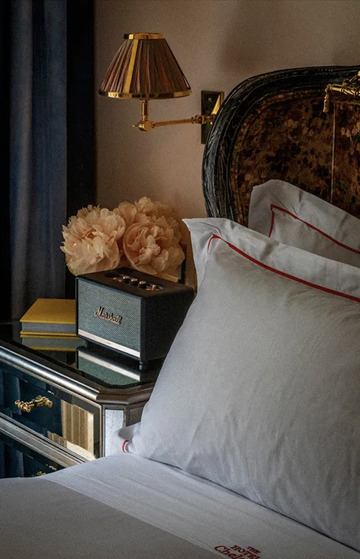 Hotel bed with a tortoiseshell headboard, monogrammed white linens, and a nightstand styled with peach roses and a vintage lamp
