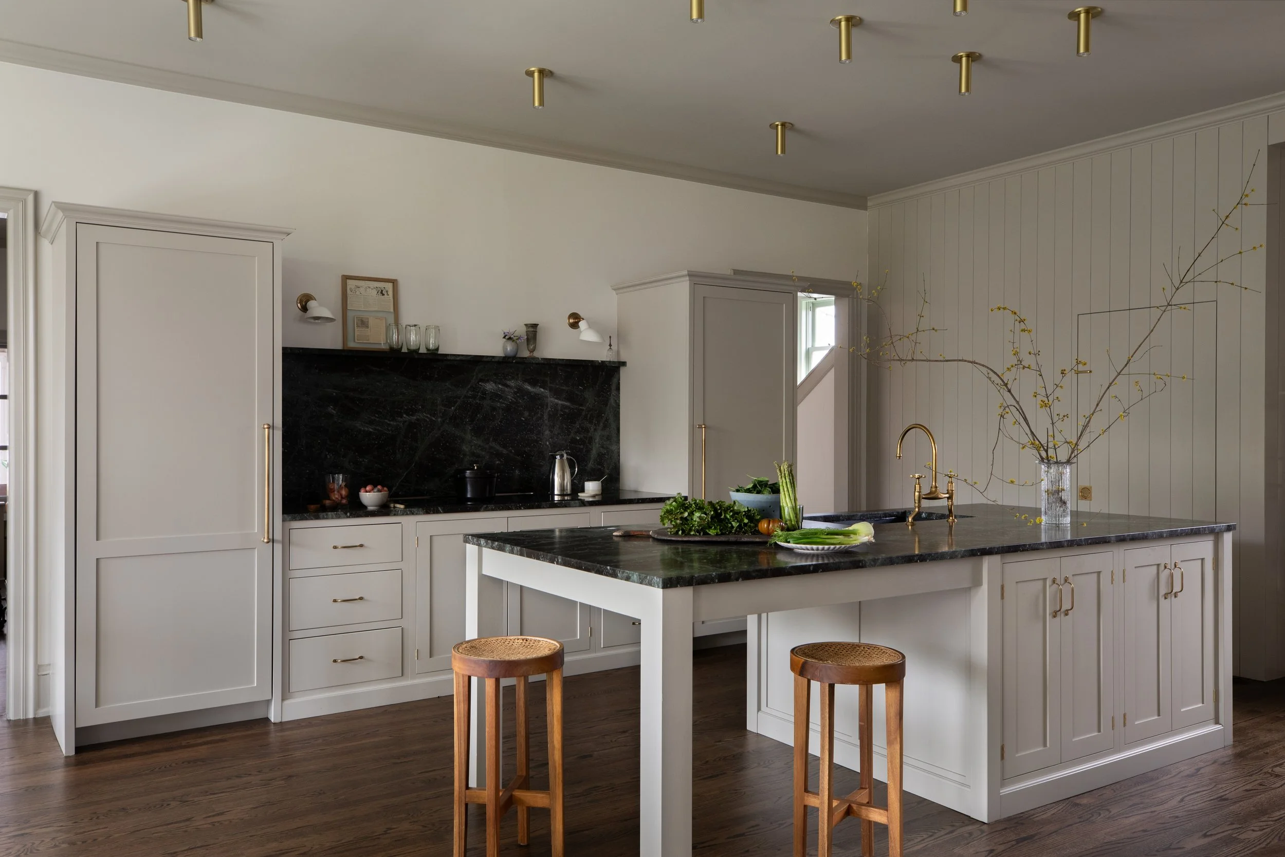 Modern kitchen with white cabinetry, black marble countertops, and a large white island with a black marble top. Two wooden bar stools are positioned at the island. A glass vase with branches and yellow buds is on the island. The kitchen has wooden flooring and a black marble backsplash with kitchen utensils and decor on it.