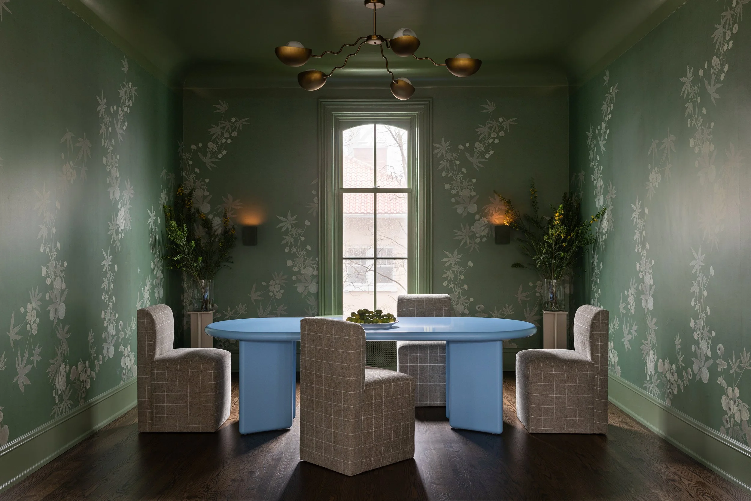 A dining room with green floral wallpaper, a large window, a blue oval table, six upholstered chairs, and a modern chandelier.