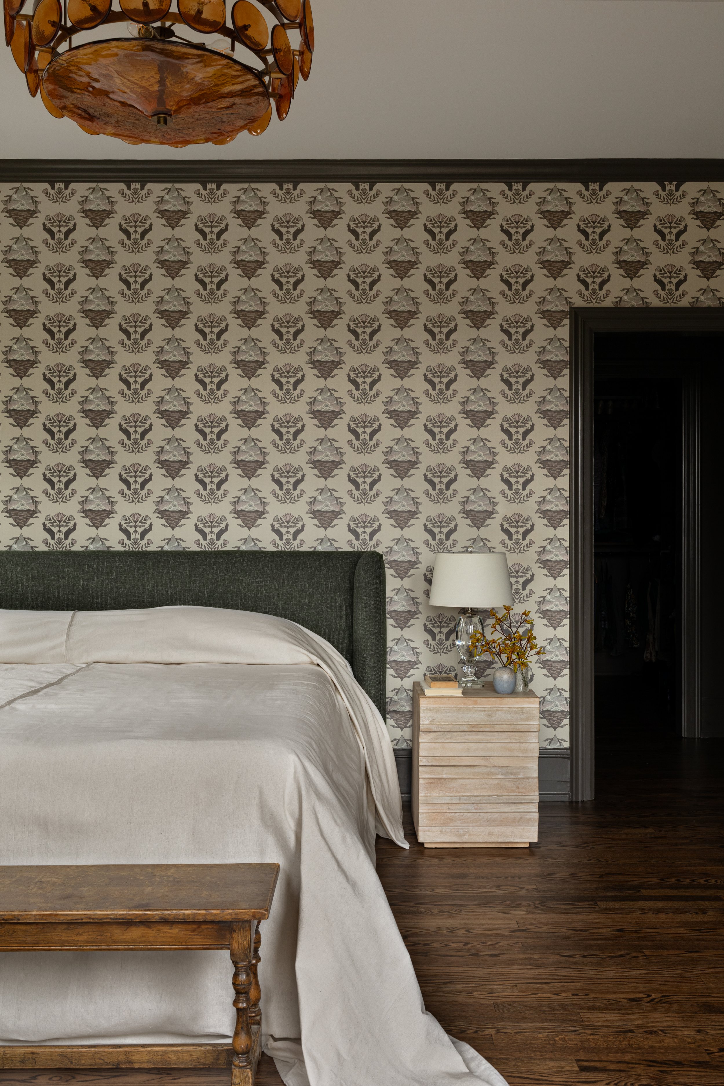 Bedroom interior featuring a bed with a dark green upholstered headboard, white bedding, and a wooden bench at the foot. A nightstand with a white lamp, decorative vase, and books. Patterned wallpaper with floral motifs, dark window trim, and a hardwood floor.