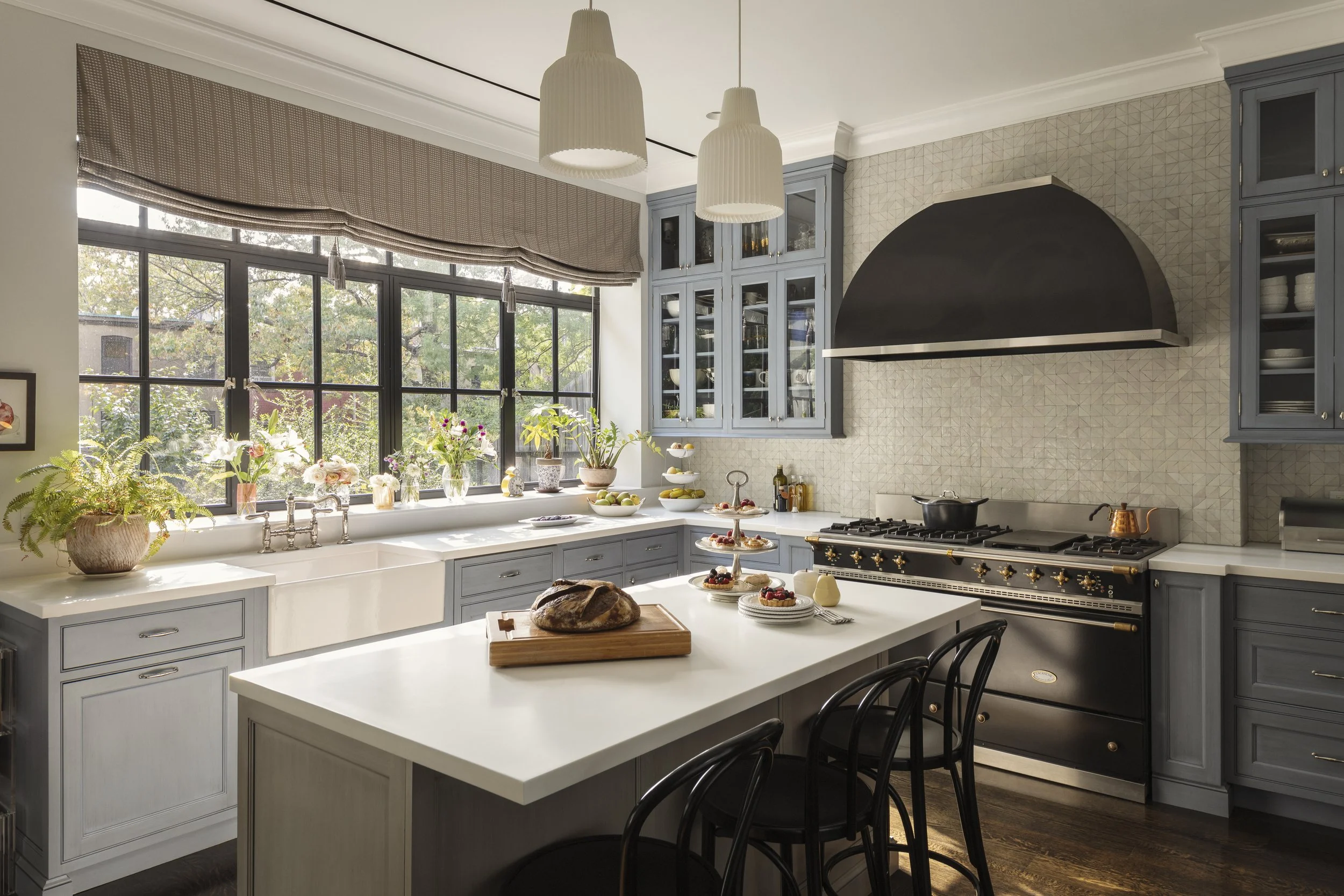 Bright kitchen with large window, white and blue cabinets, and black stove with black range hood. Countertop has plants, baked bread, desserts, and bowls.