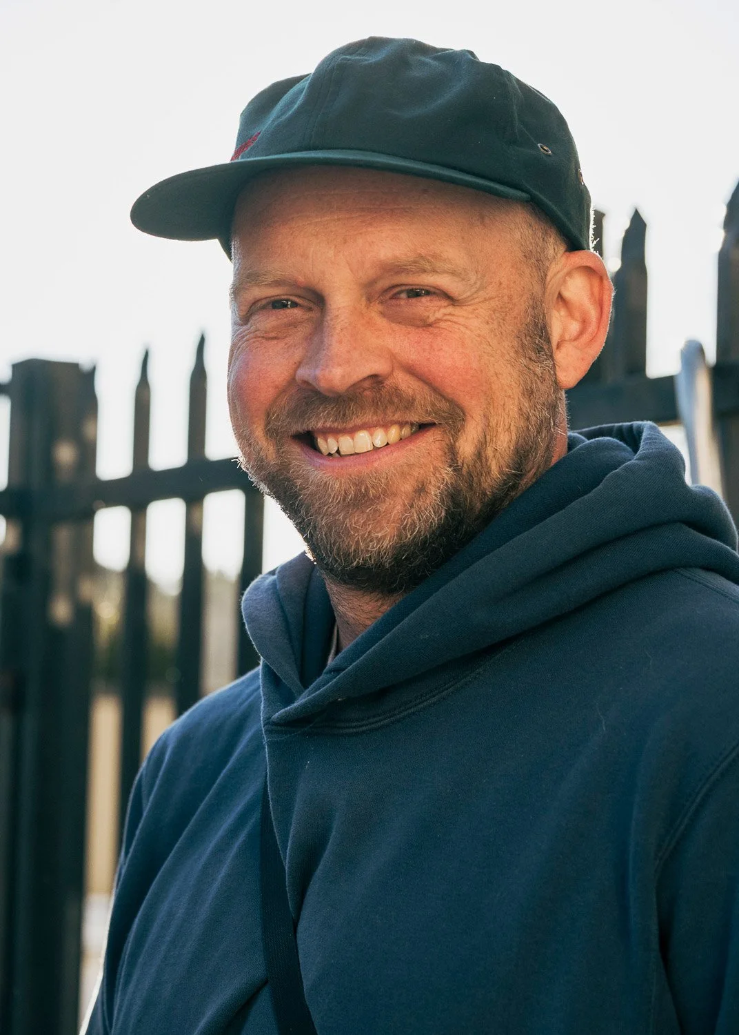 A smiling man wearing a dark cap and hoodie outdoors with a fence in the background.