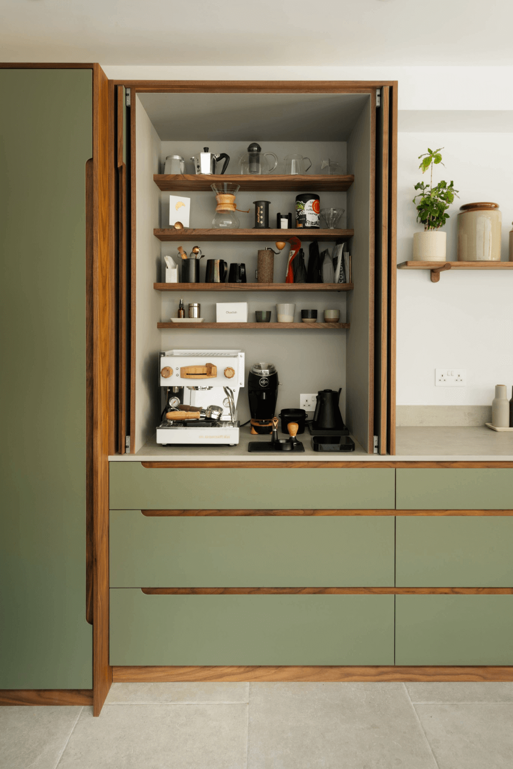 Open kitchen cabinet with coffee machine, coffee grinder, and various kitchen items on shelves and countertop, in a modern kitchen with green cabinets and minimalist decor.
