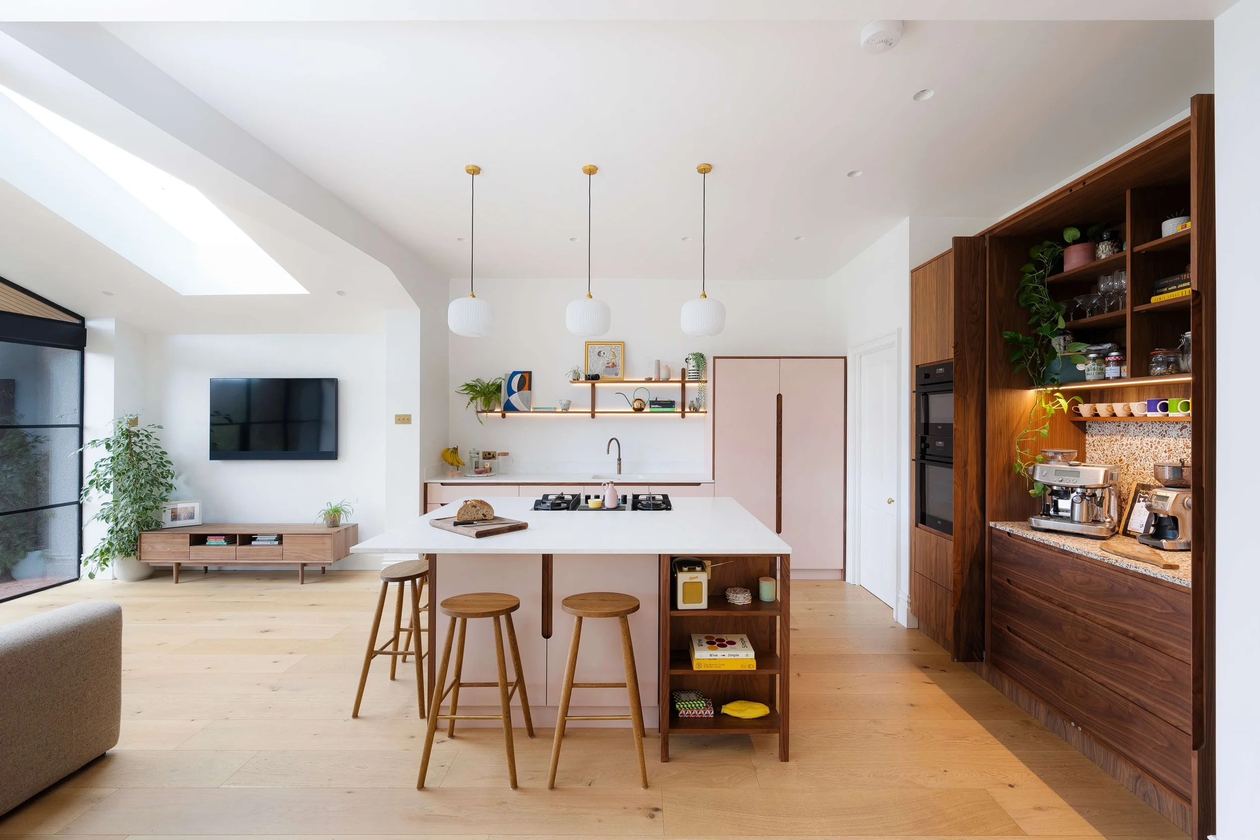 Modern open-plan kitchen with white island, wooden stools, and wooden shelving unit displaying plants, dishes, and kitchen appliances, with a living area and wall-mounted TV in the background.