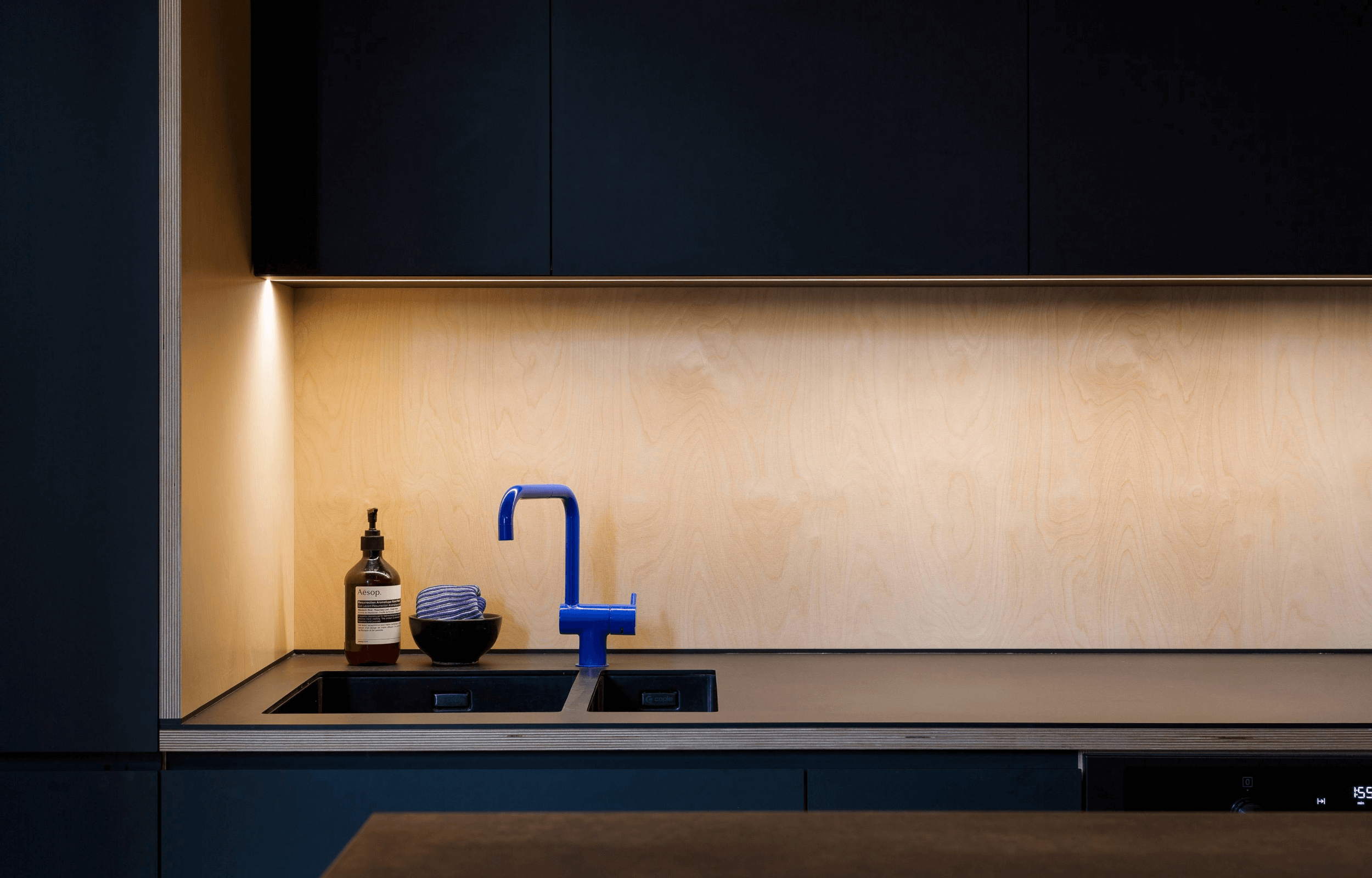 A modern kitchen with dark blue cabinets, a black countertop, a blue faucet, a bottle of Aesop hand soap, a black bowl with a striped cloth, and warm lighting on a wooden backsplash.