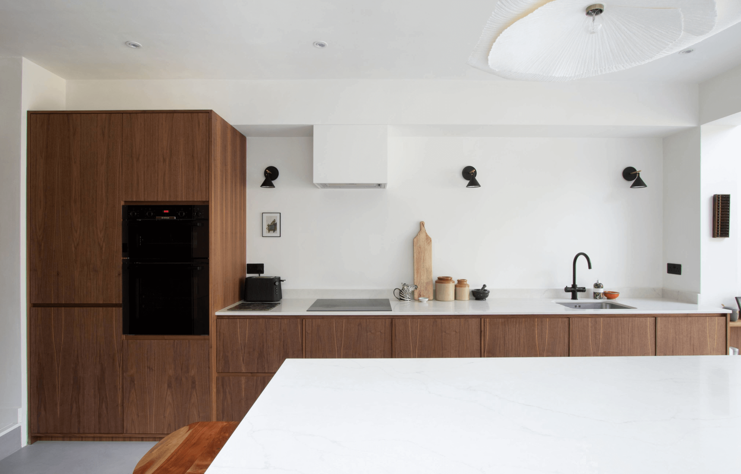 Modern kitchen with white countertop, dark wood cabinets, black built-in oven, small black appliances, and various kitchen utensils on the counter.